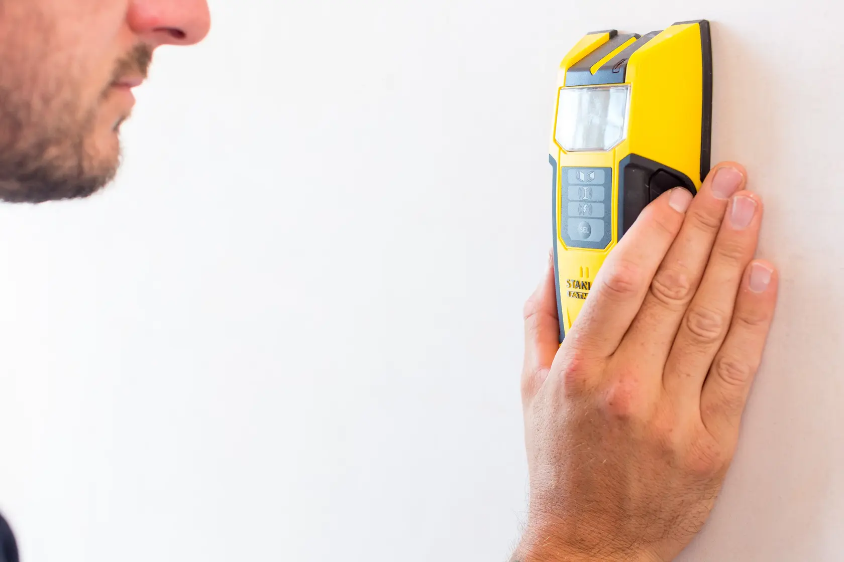 STANLEY FMHT0-77407 stud finder in yellow held against a white wall by a hand, front angle.