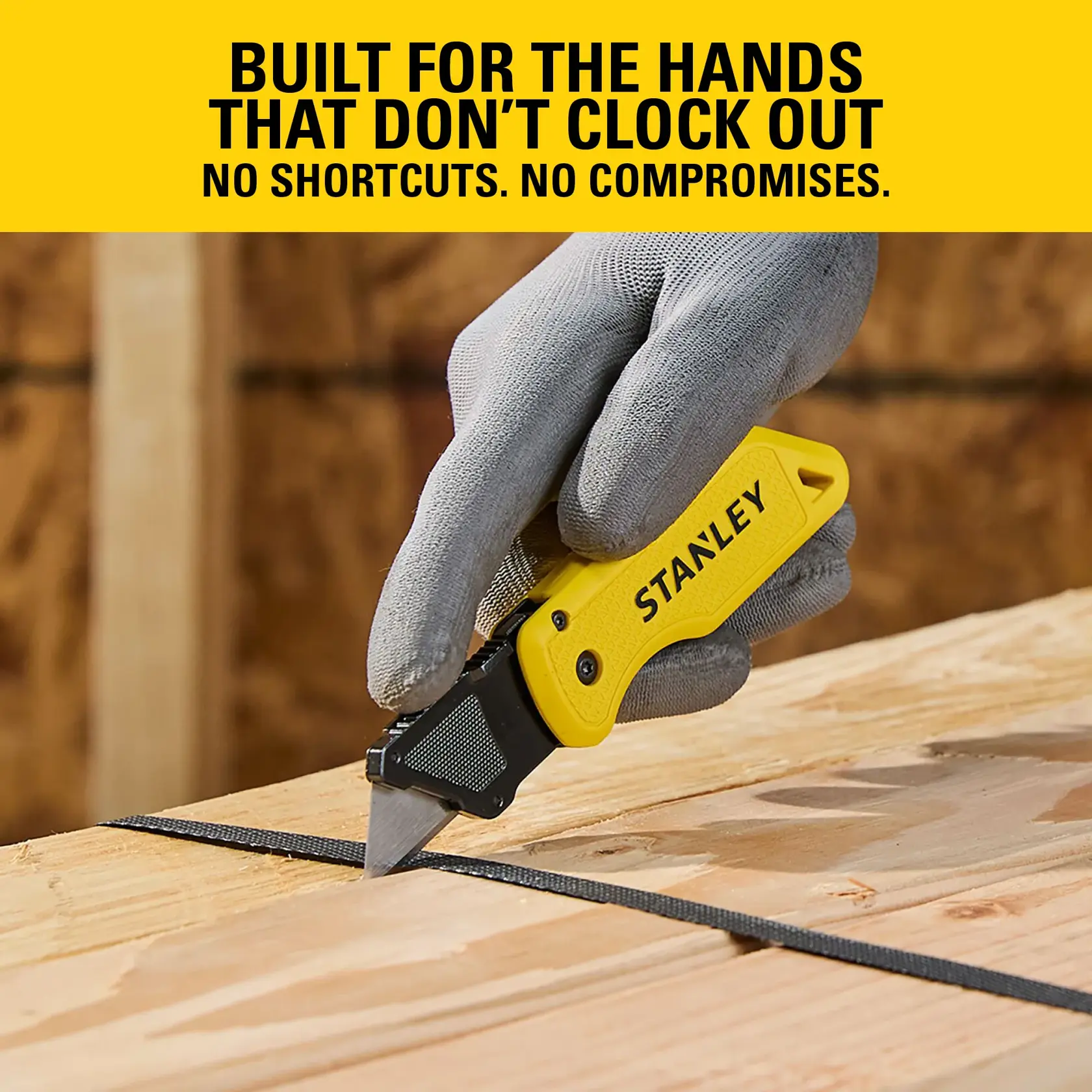 Stanley utility knife with yellow handle, used to cut black strap on wooden surface, hand in gray glove.