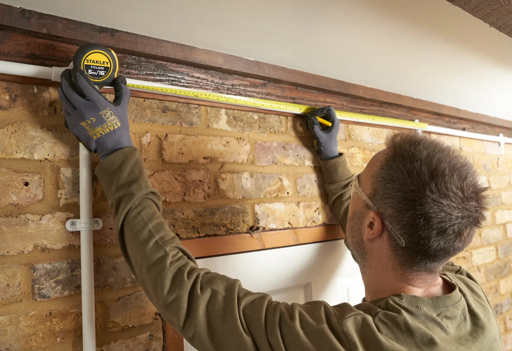 Person measuring wall above door with STANLEY 0-30-696 tape measure, brick background, indoor setting.