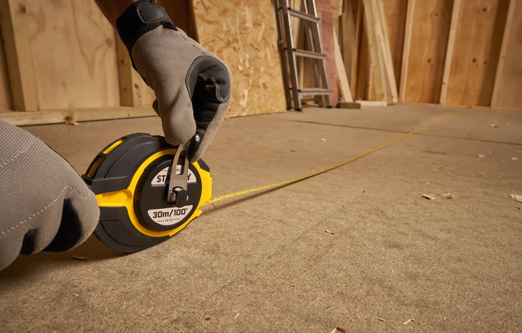 Photo of STANLEY 0-34-132 long tape measure in use on construction floor, yellow and black casing, gloved hand visible.