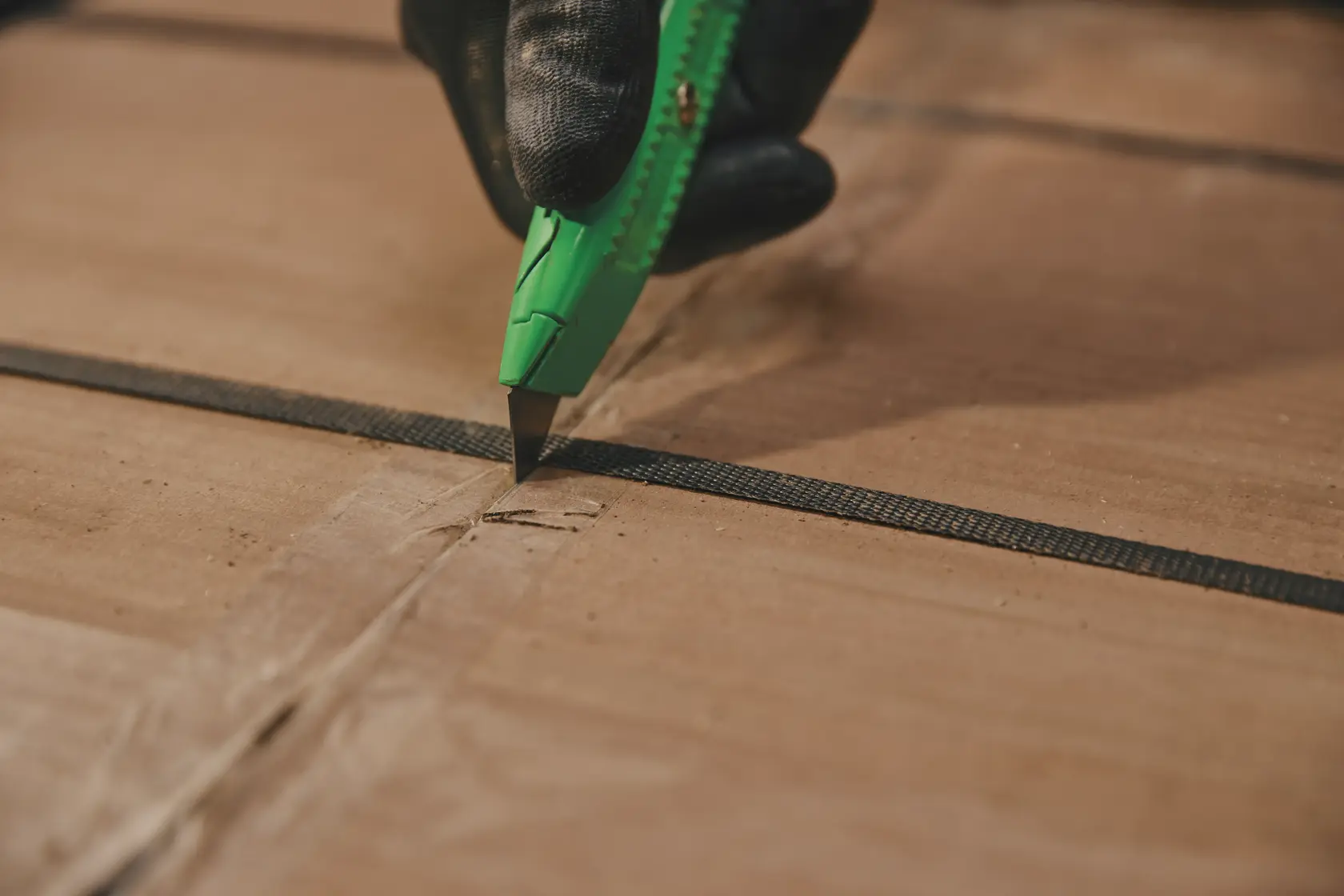 Photo of STANLEY 10-179 utility knife cutting cardboard box, close-up view of green handle and gloved hand.