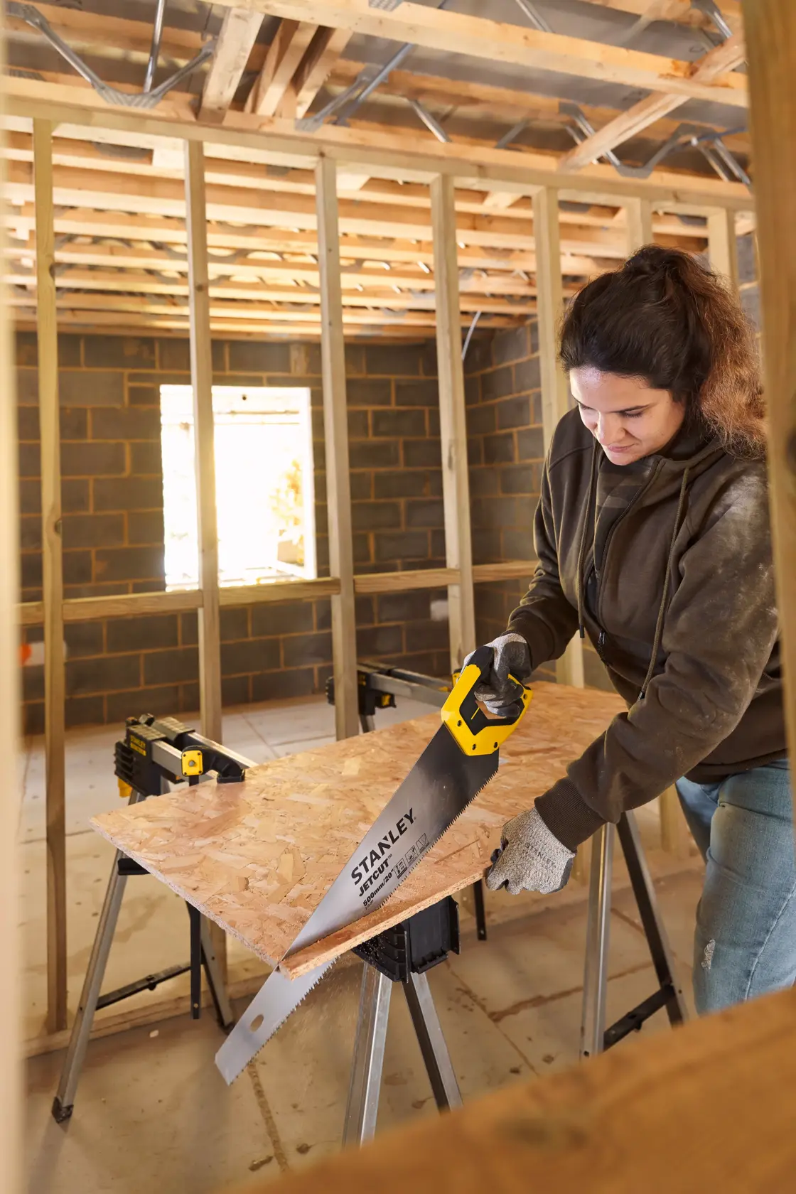 STANLEY 2-15-288 hand saw with yellow handle cutting wood board on sawhorses in construction site, side view.