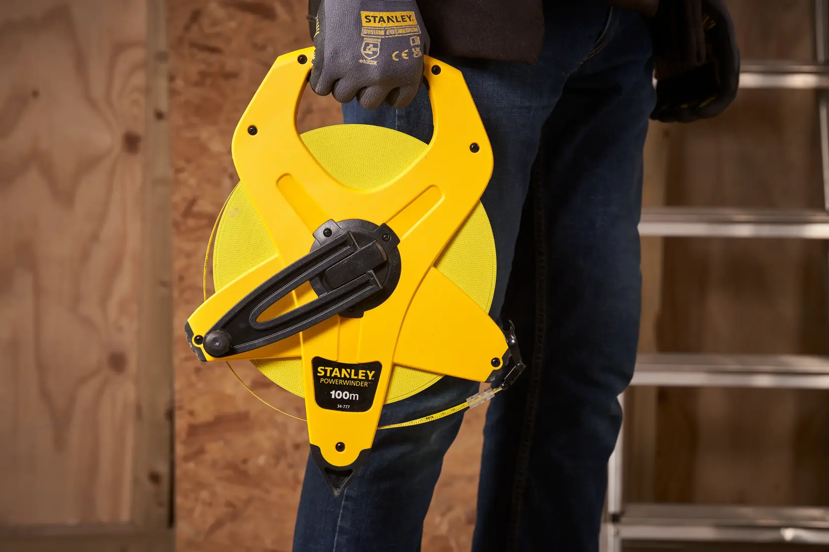 STANLEY 2-34-777 open reel measuring tape held by person wearing gloves, construction site background.