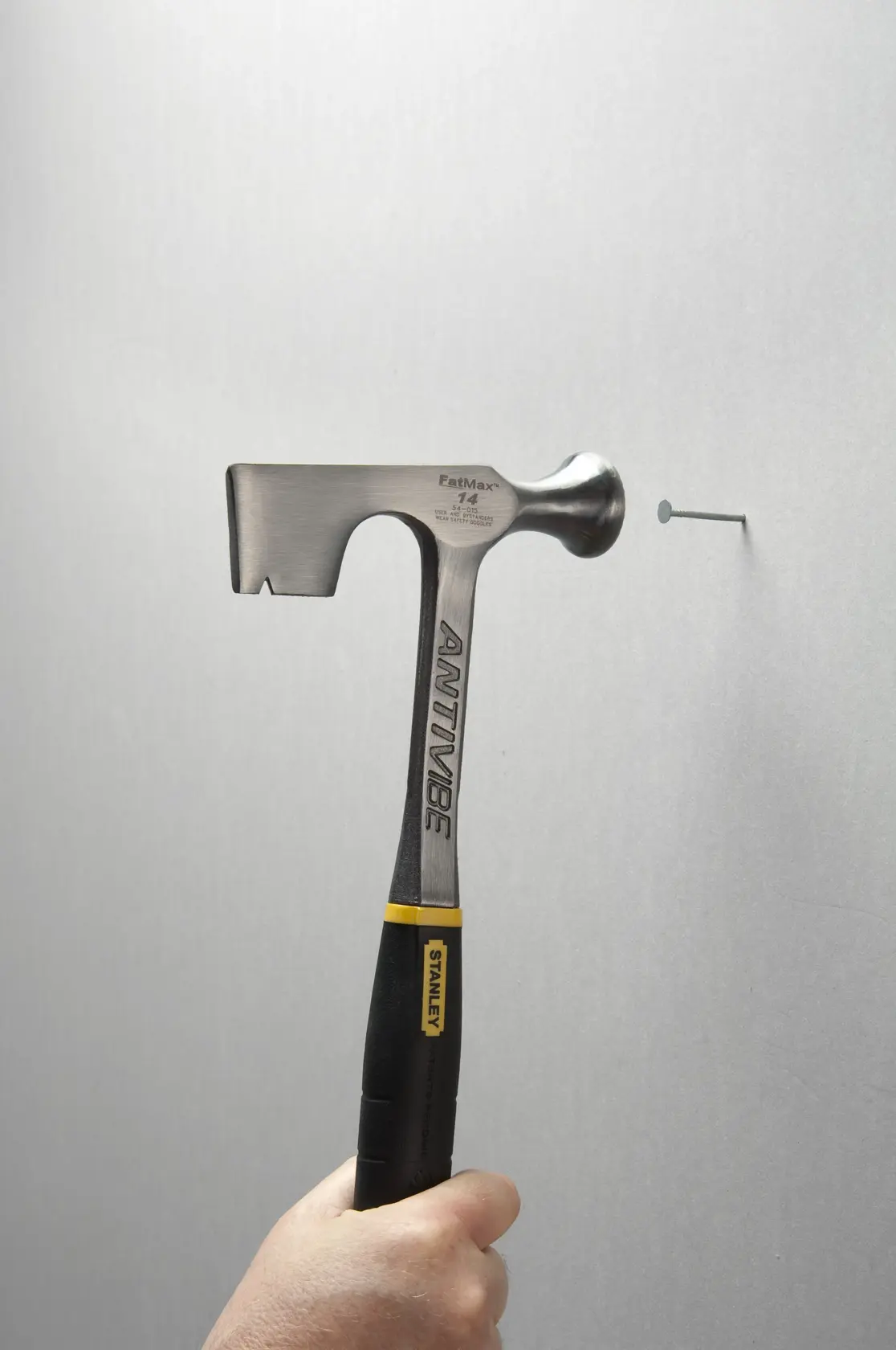 Photo of STANLEY 54-015 hammer with black grip striking a nail against a gray wall, angled side view.