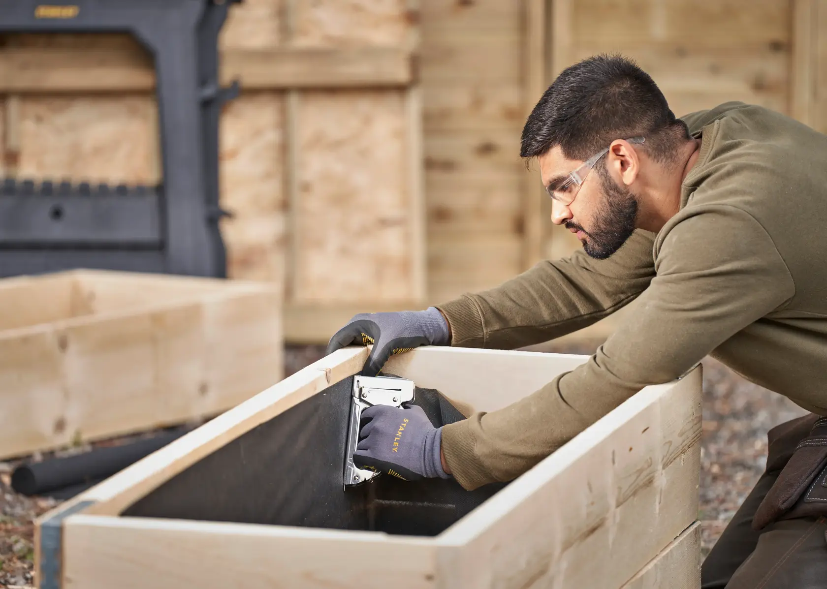 STANLEY 6-TR45 staple gun used by a person wearing gloves to assemble a wooden planter, outdoor setting.