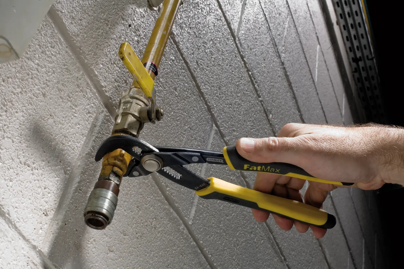A person uses a pair of Stanley FatMax adjustable pliers to grip and turn a pipe fitting attached to a yellow pipe against a textured white wall.