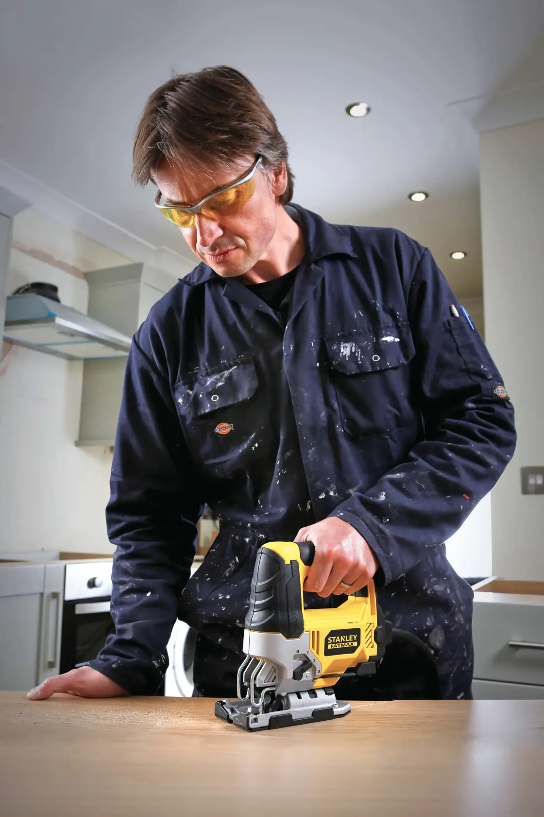STANLEY FME340K jigsaw held by person in blue coveralls cutting wood on workbench in kitchen setting.