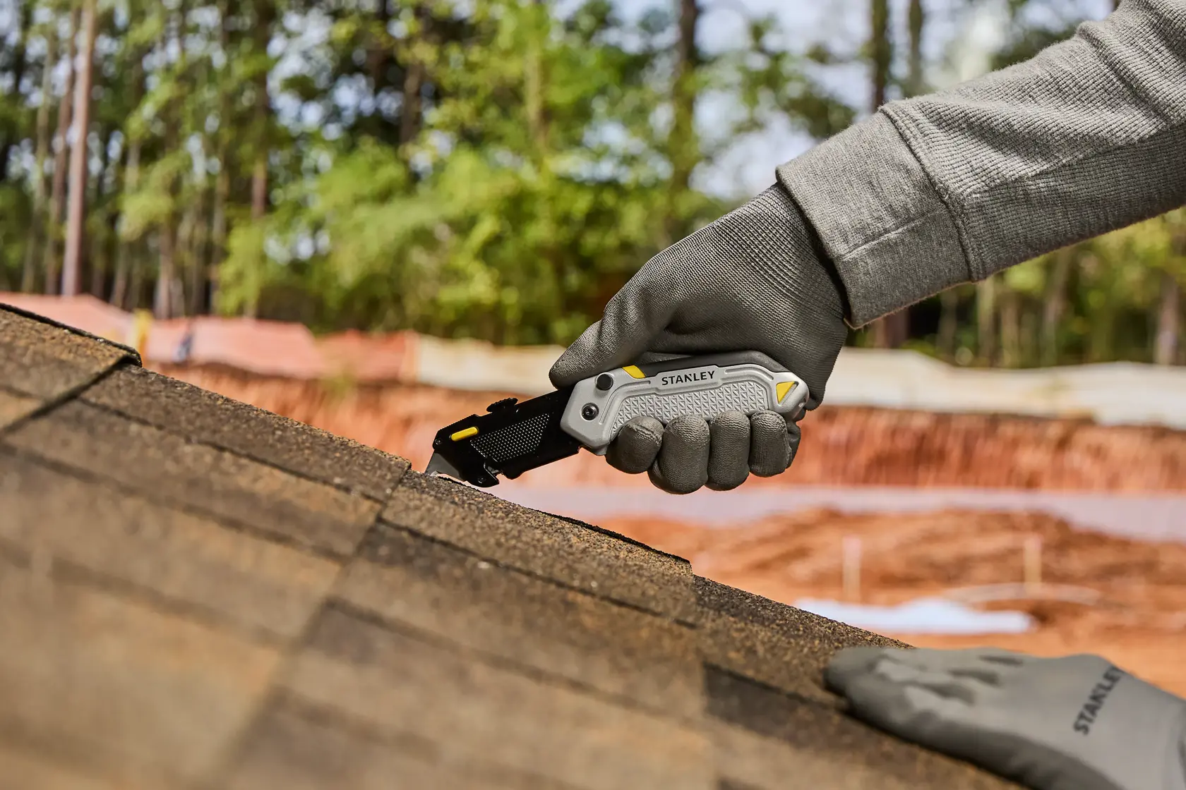 A person wearing work gloves uses a STANLEY utility knife to cut roofing shingles outdoors on a sloped roof.