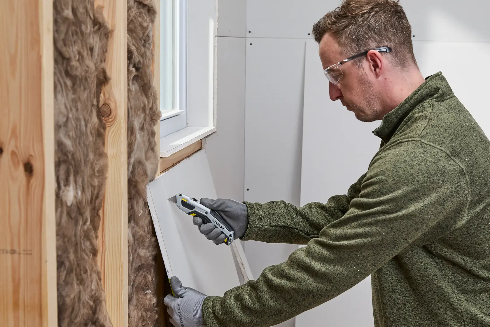 A person wearing gloves and a green sweater is using a Stanley utility knife to cut drywall near a window. The wall includes wooden studs and insulation material.