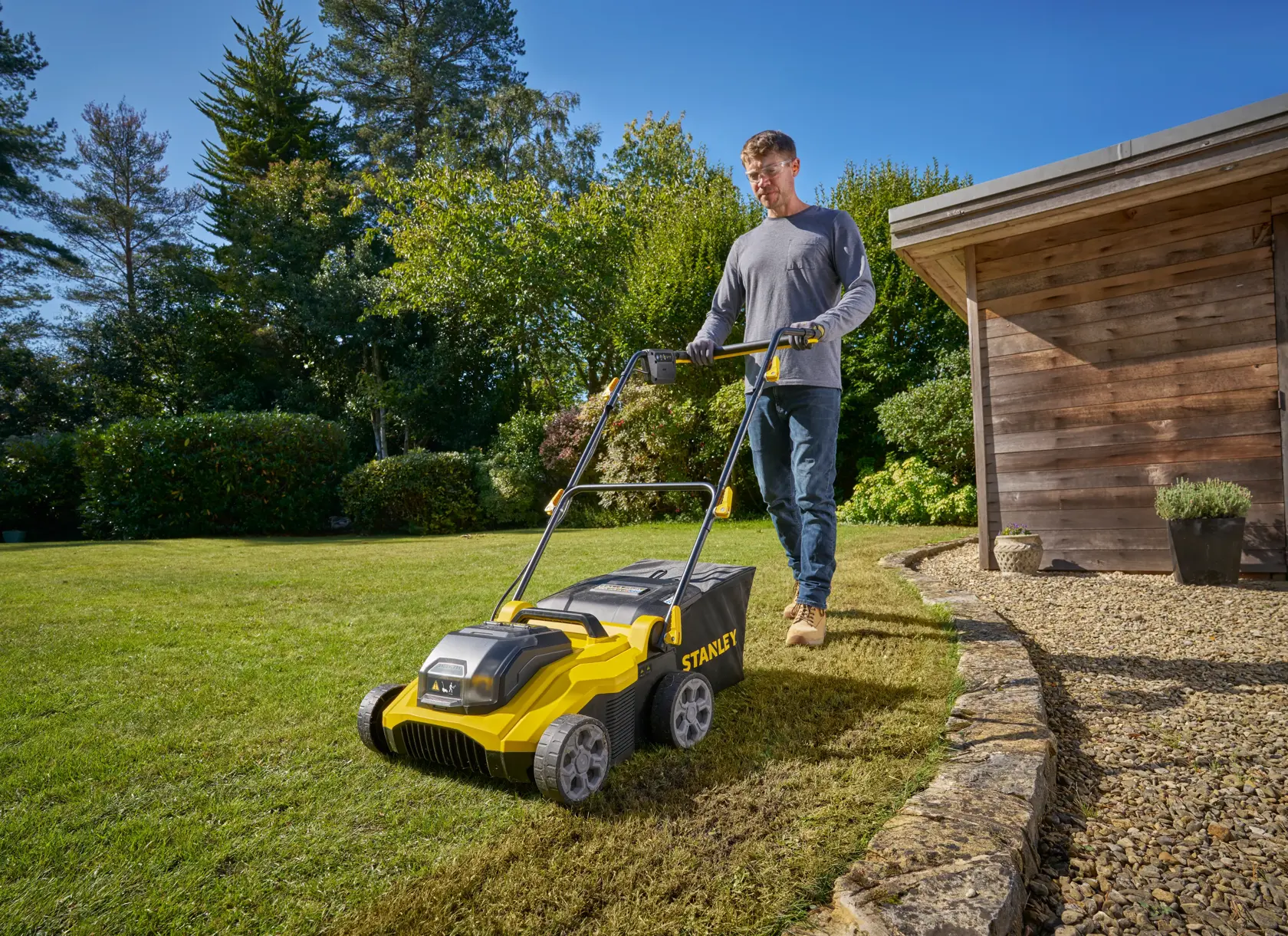 STANLEY SCOEP3801N lawn scarifier in use on a garden lawn, outdoor angle with greenery and shed.