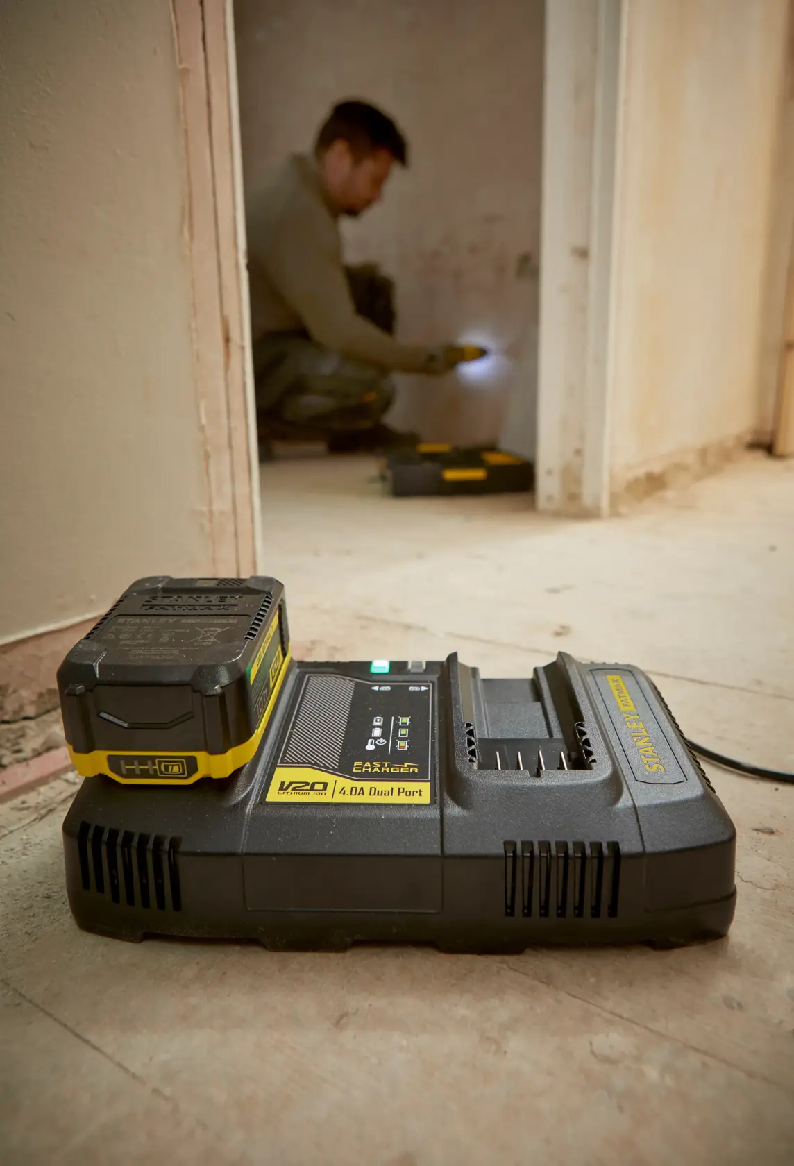 STANLEY SFMCB24 V20 dual port battery charger on workshop floor with battery attached, worker in background.