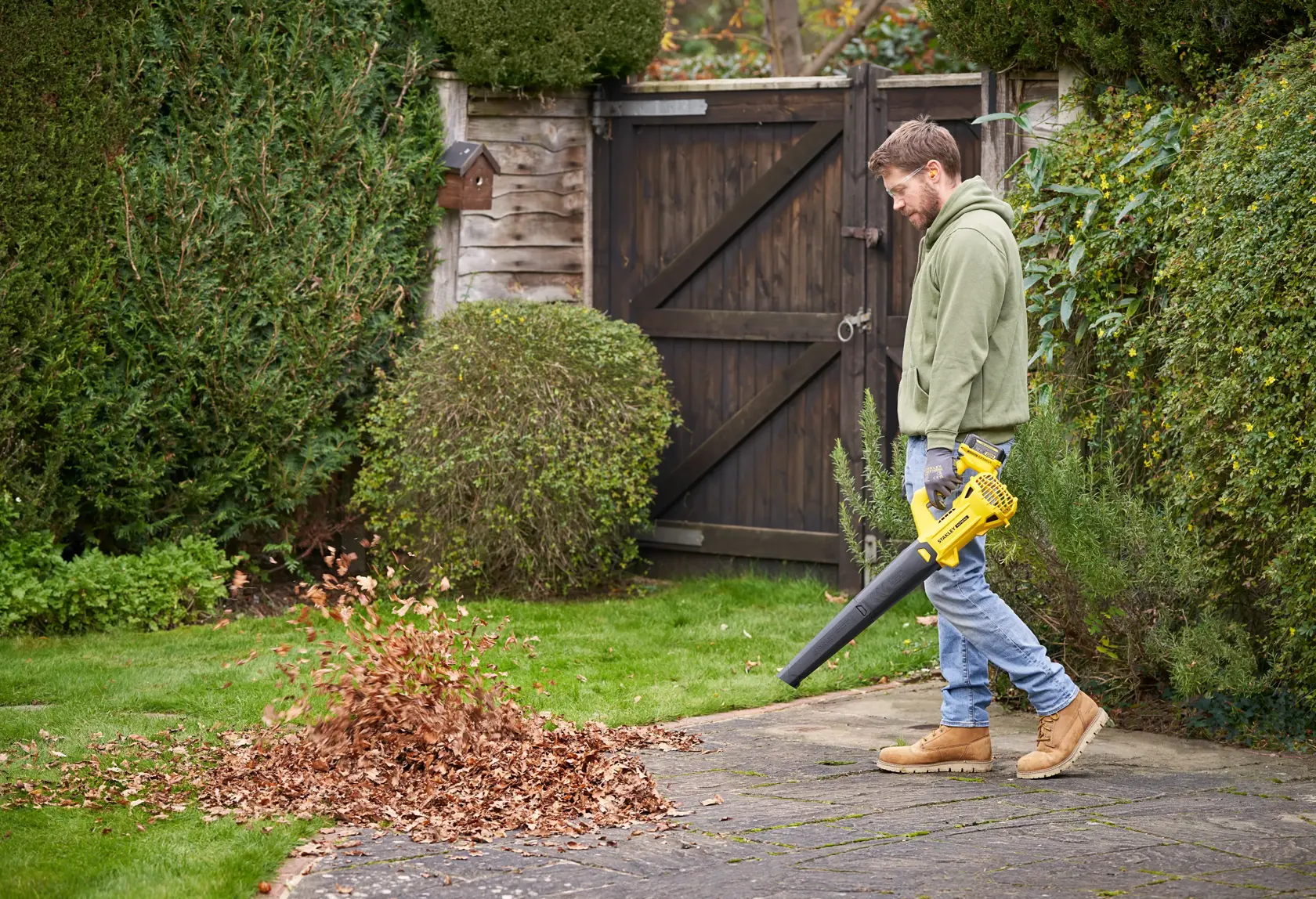 Photo of person using STANLEY SFMCBL714M1 cordless leaf blower to clear leaves from garden patio.