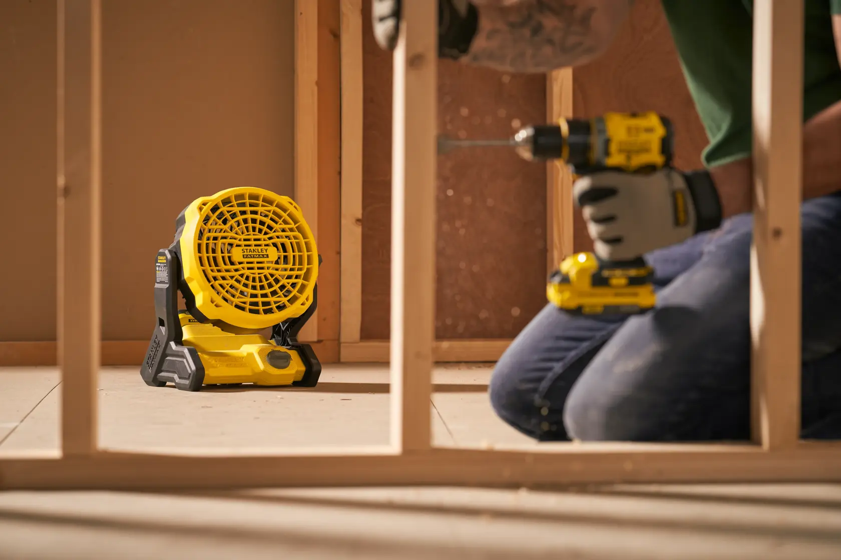 Stanley SFMCE001B portable jobsite fan on floor in construction setting, worker using power drill nearby.