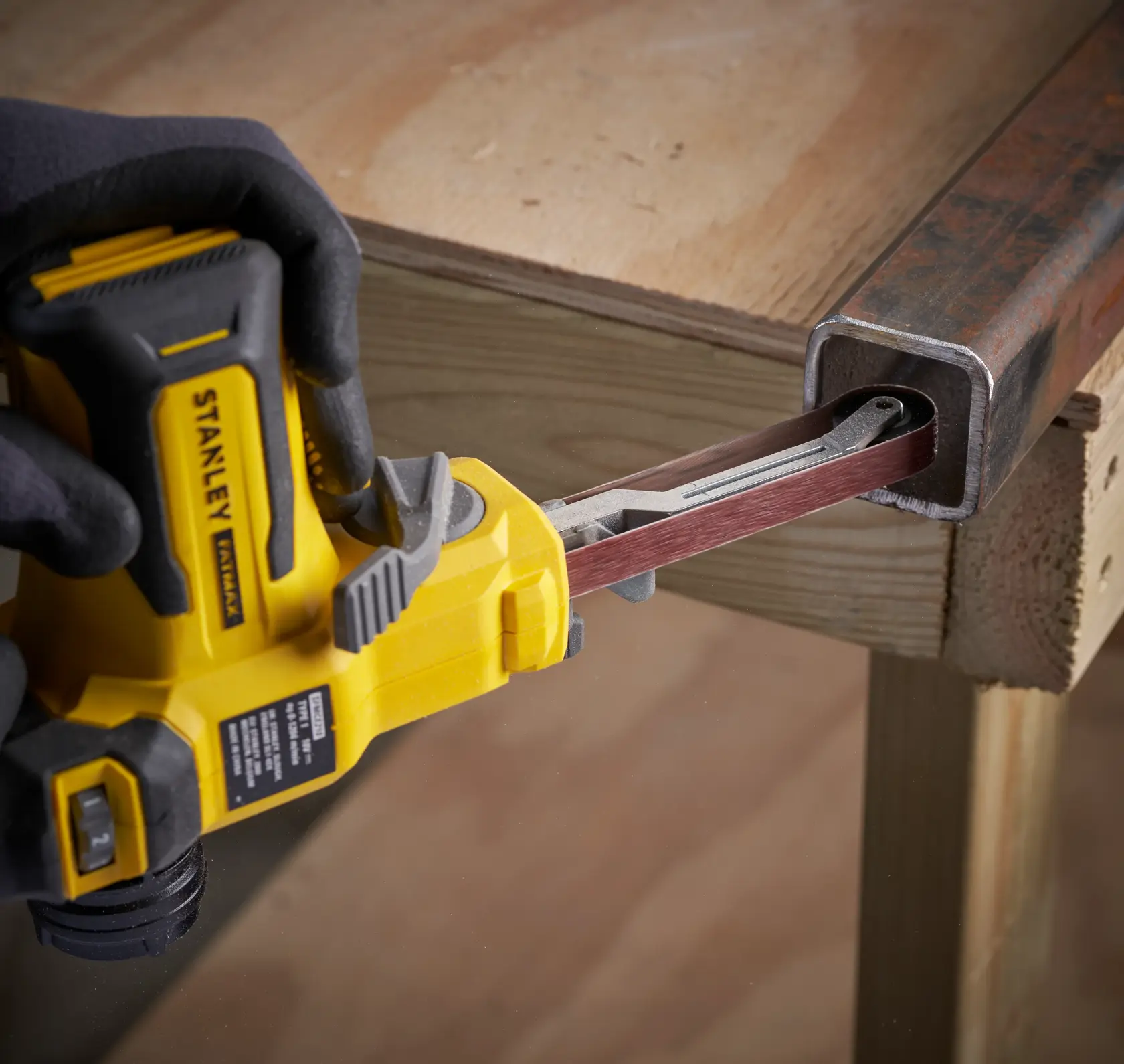 STANLEY SFMCE210B cordless belt file sander smoothing inside metal pipe, side angle, held by gloved hand.