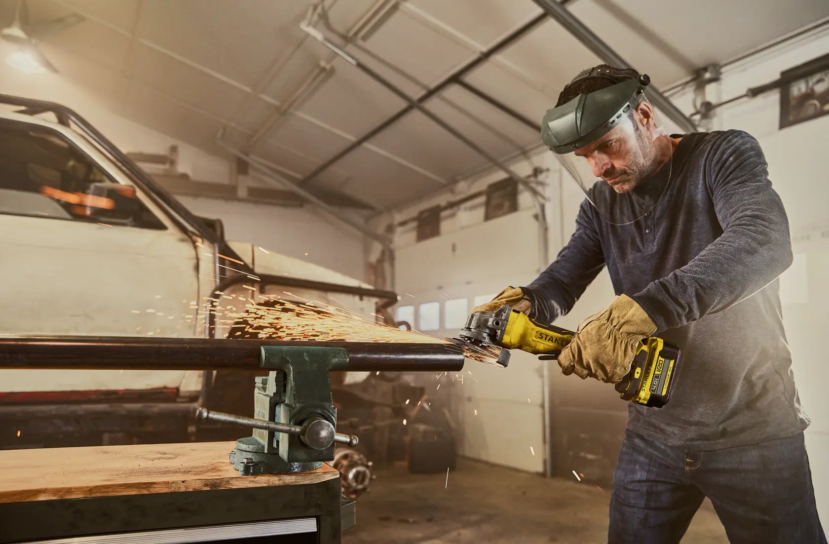 STANLEY SFMCG700B cordless angle grinder in use, producing sparks as it grinds metal pipe in a workshop.