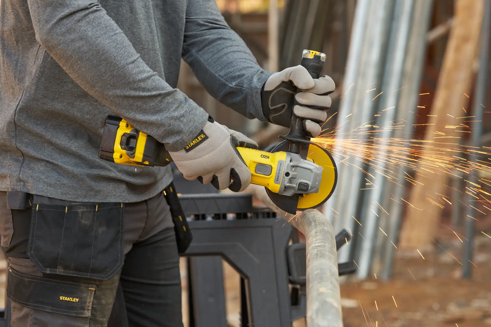 Stanley SFMCG710B-XJ cordless angle grinder in use cutting metal pipe, yellow and black tool, sparks visible.