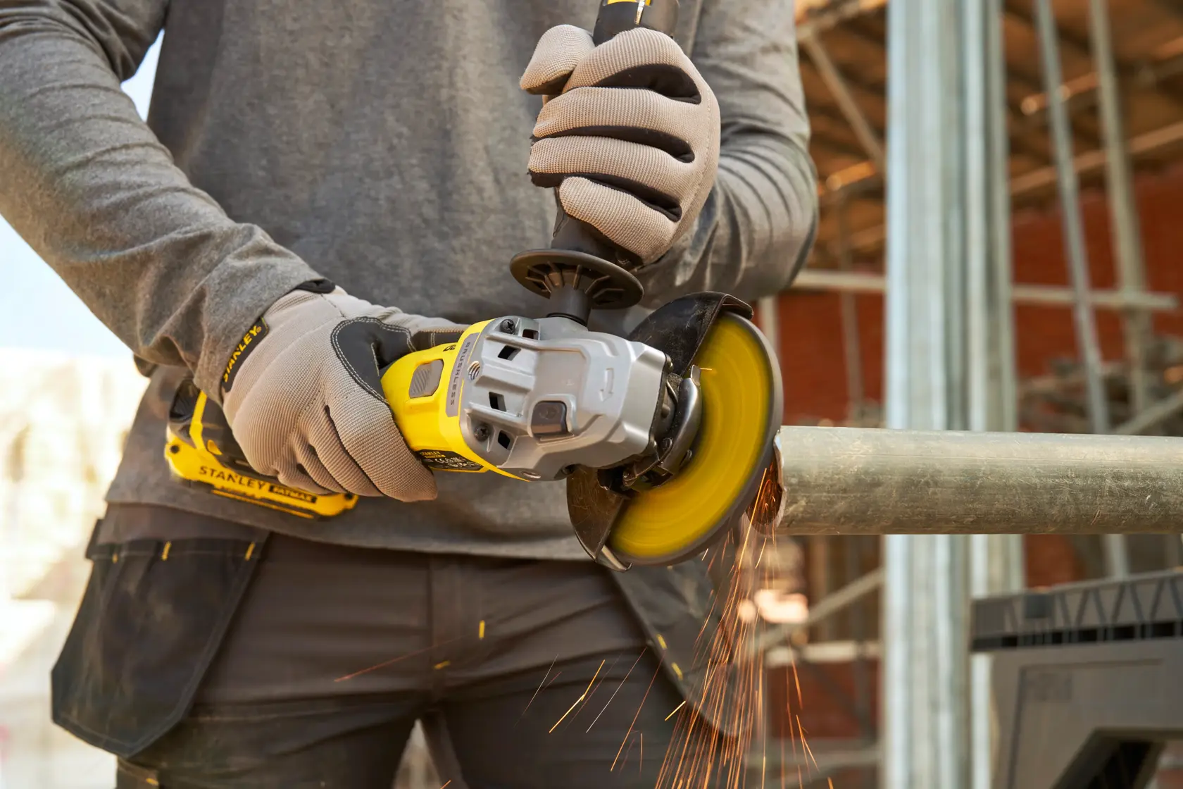 Photo of worker using STANLEY SFMCG710B-XJ cordless angle grinder cutting metal pipe, sparks flying, workshop setting.