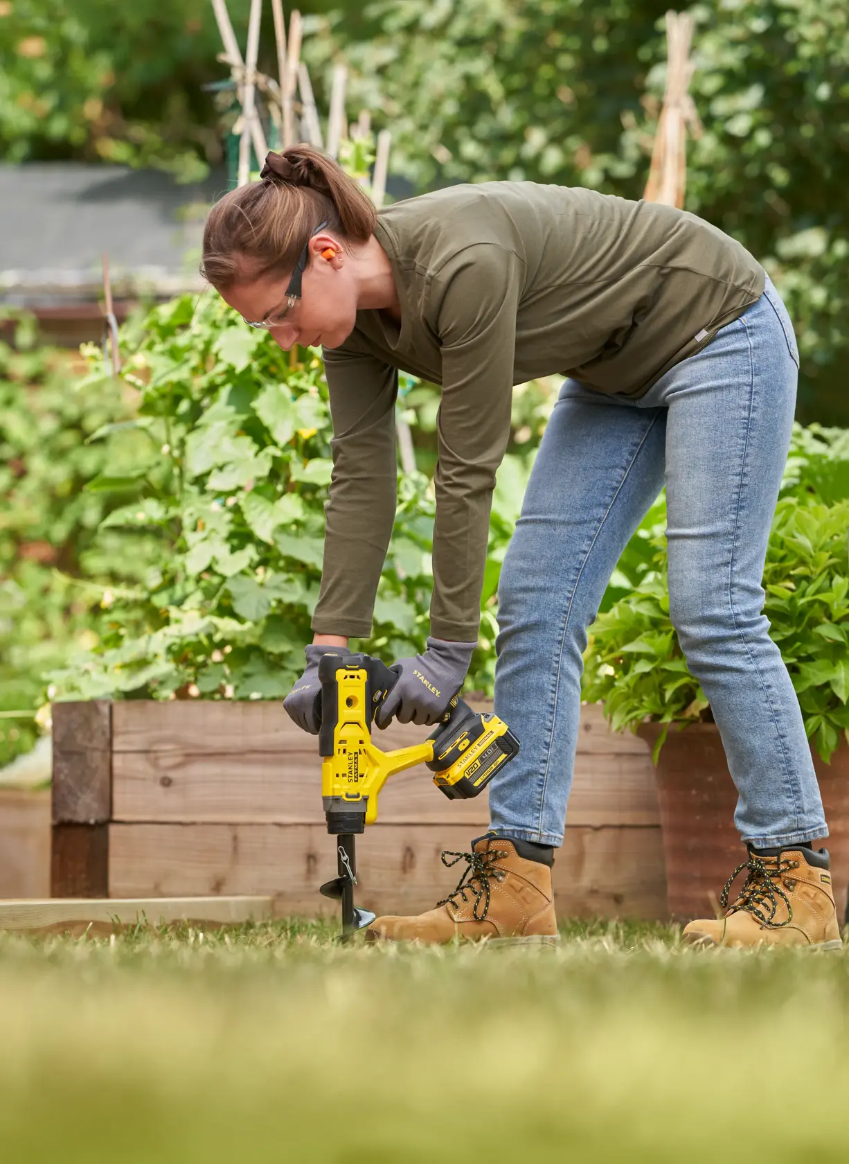 Photo of person using STANLEY SFMCGA75B cordless earth auger in a garden, drilling into soil near raised beds.