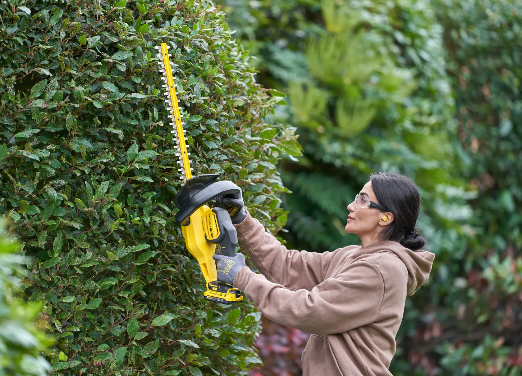 STANLEY SFMCHT650D1 hedge trimmer in use, yellow and black, trimming green bushes outdoors.