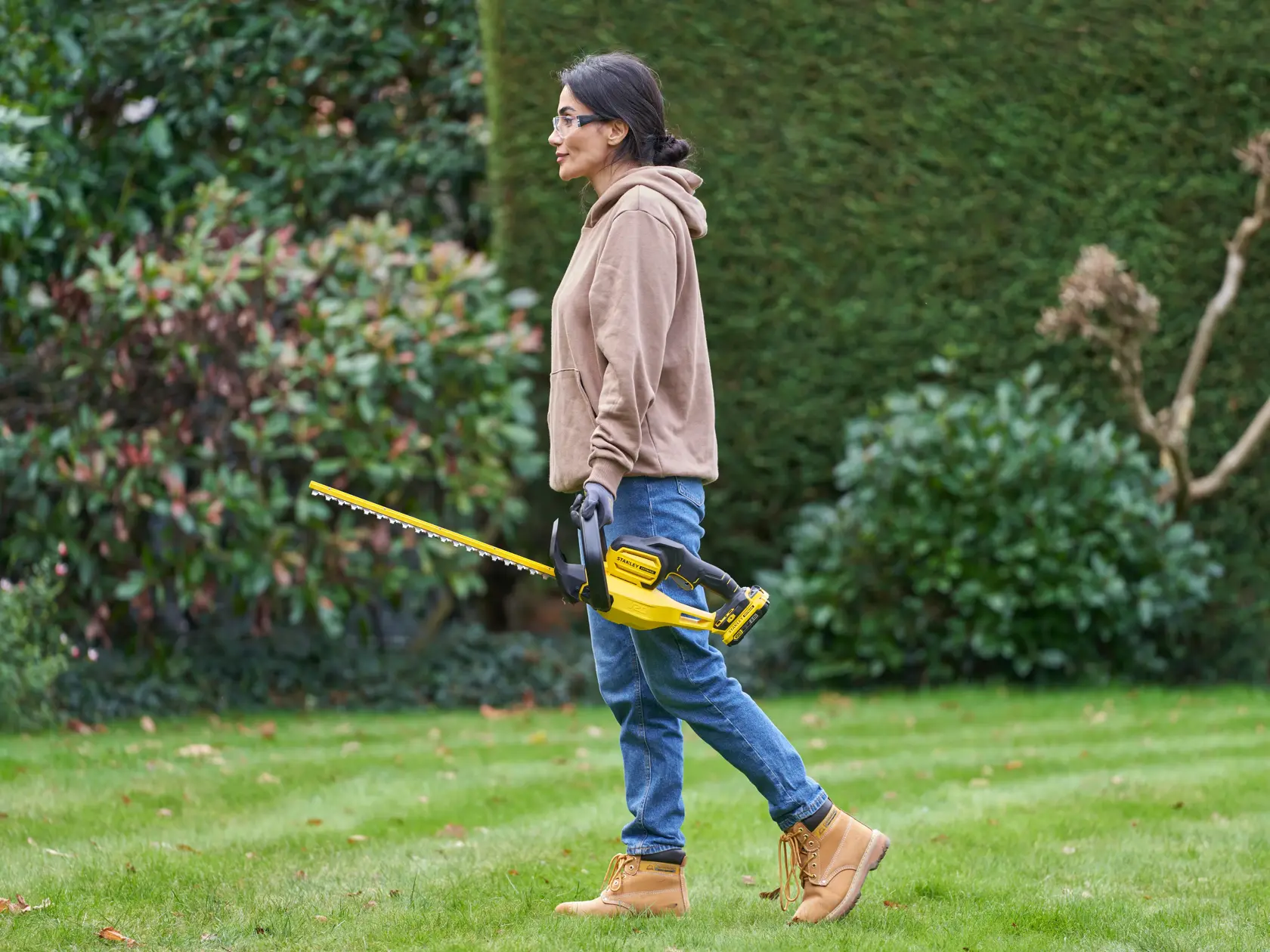 STANLEY SFMCHT650D1 hedge trimmer in use outdoors, person holding yellow and black tool near green bushes.