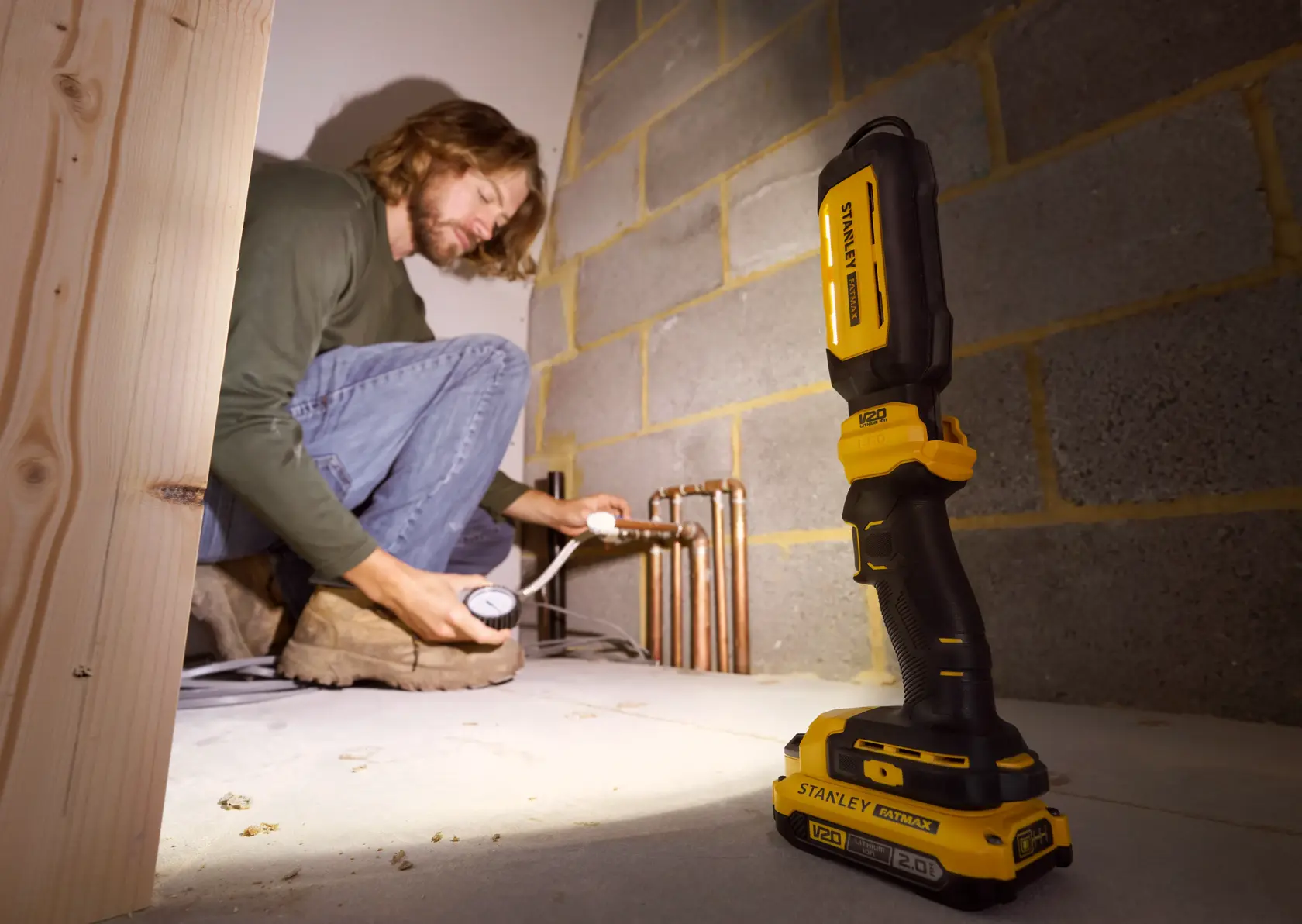 STANLEY SFMCL050B 18V FATMAX V20 Pivot Light on floor, illuminating pipes, person working in background.