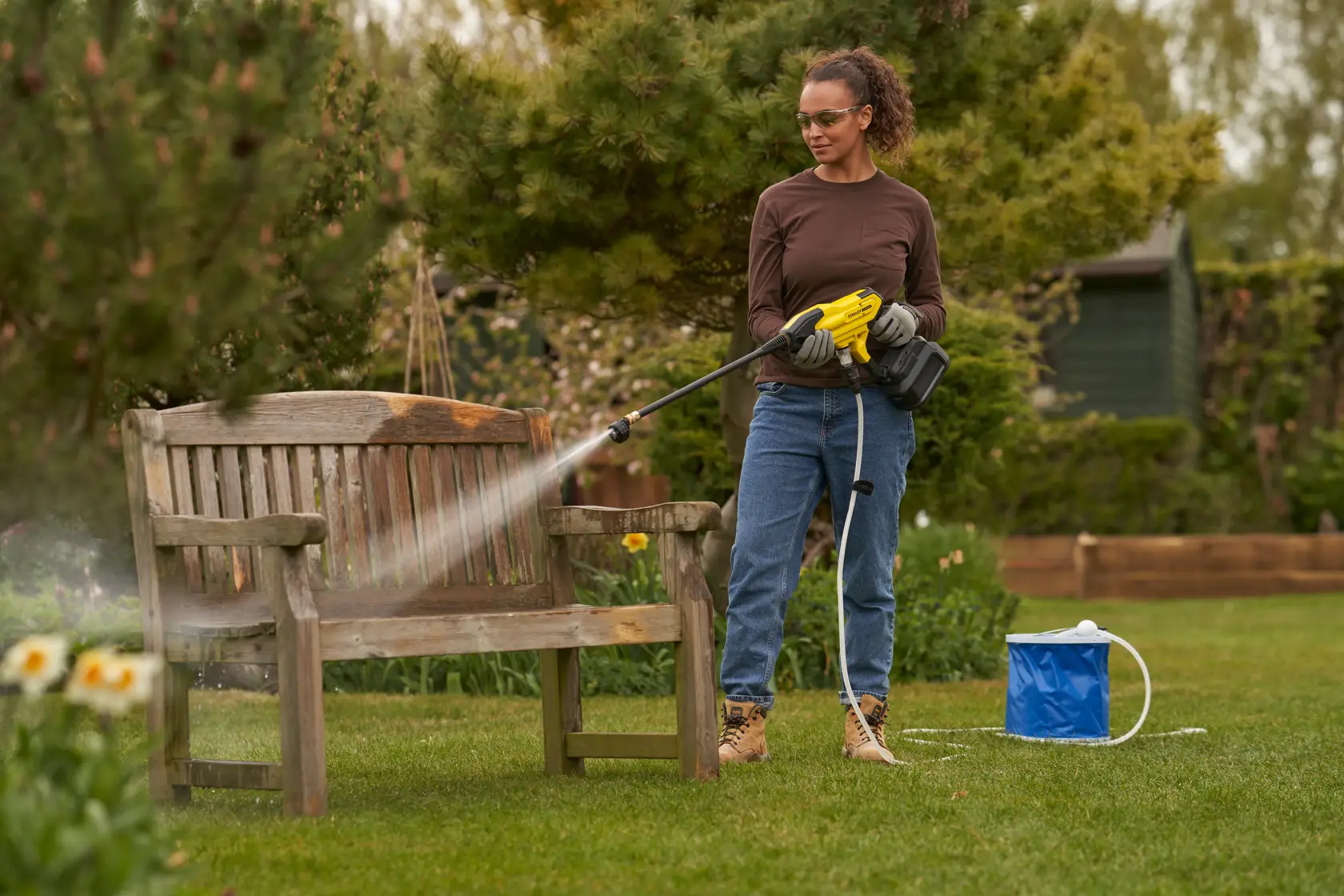 STANLEY SFMCPC93B portable pressure washer spraying wooden bench outdoors, with blue water container on grass.