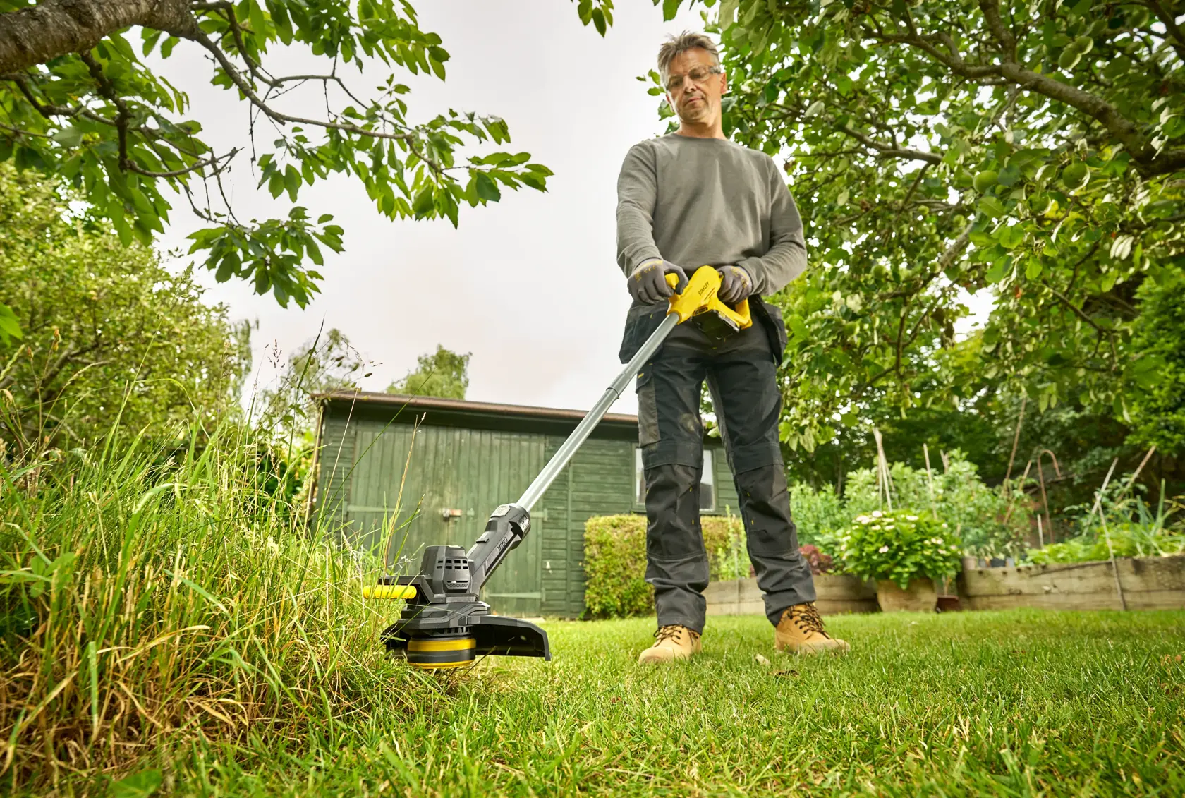 STANLEY SFMCST925M1 grass trimmer in use, person trimming lawn in garden with green shed in background.