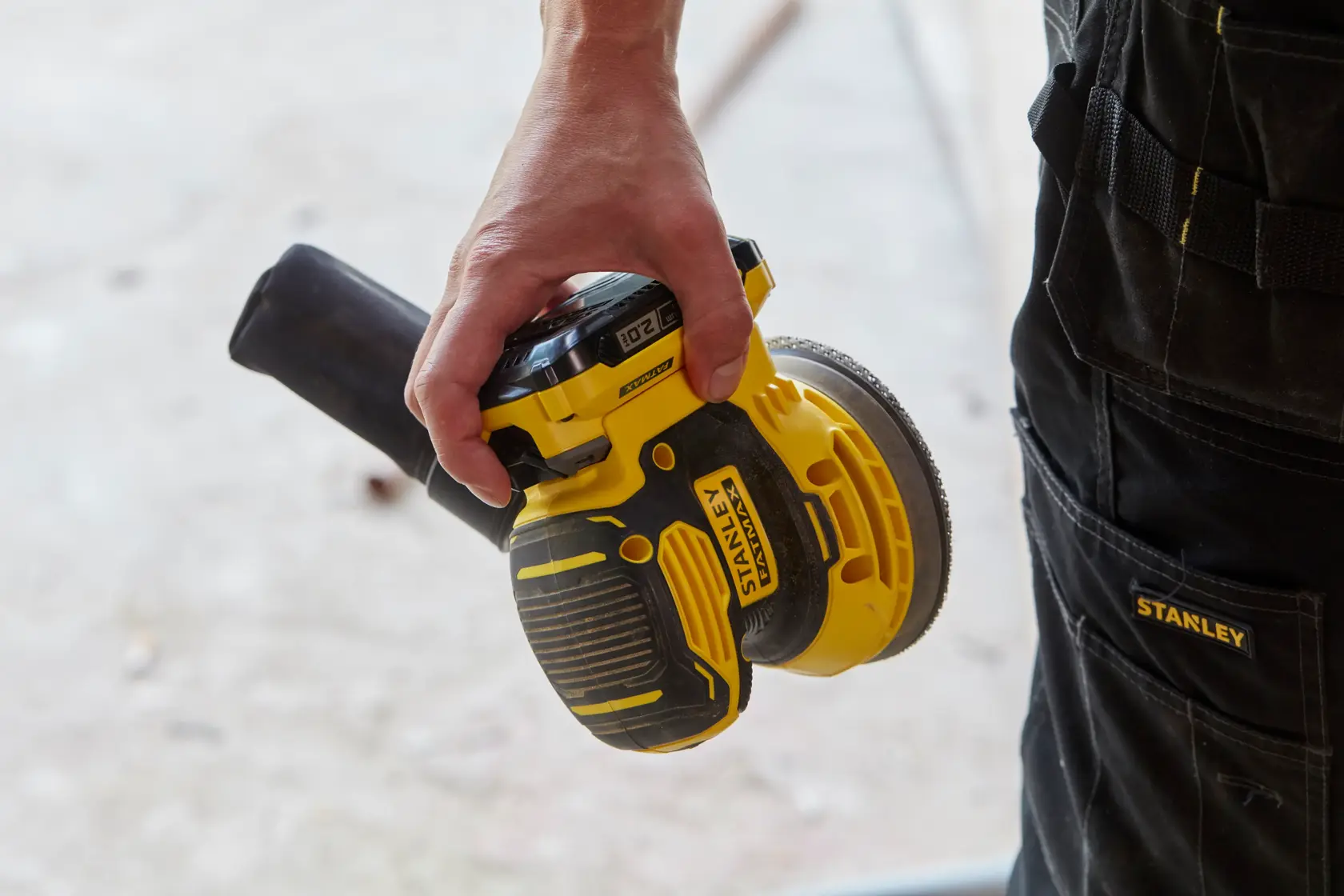 Photo of STANLEY SFMCW220B cordless orbital sander in hand, yellow and black body, work pants visible.