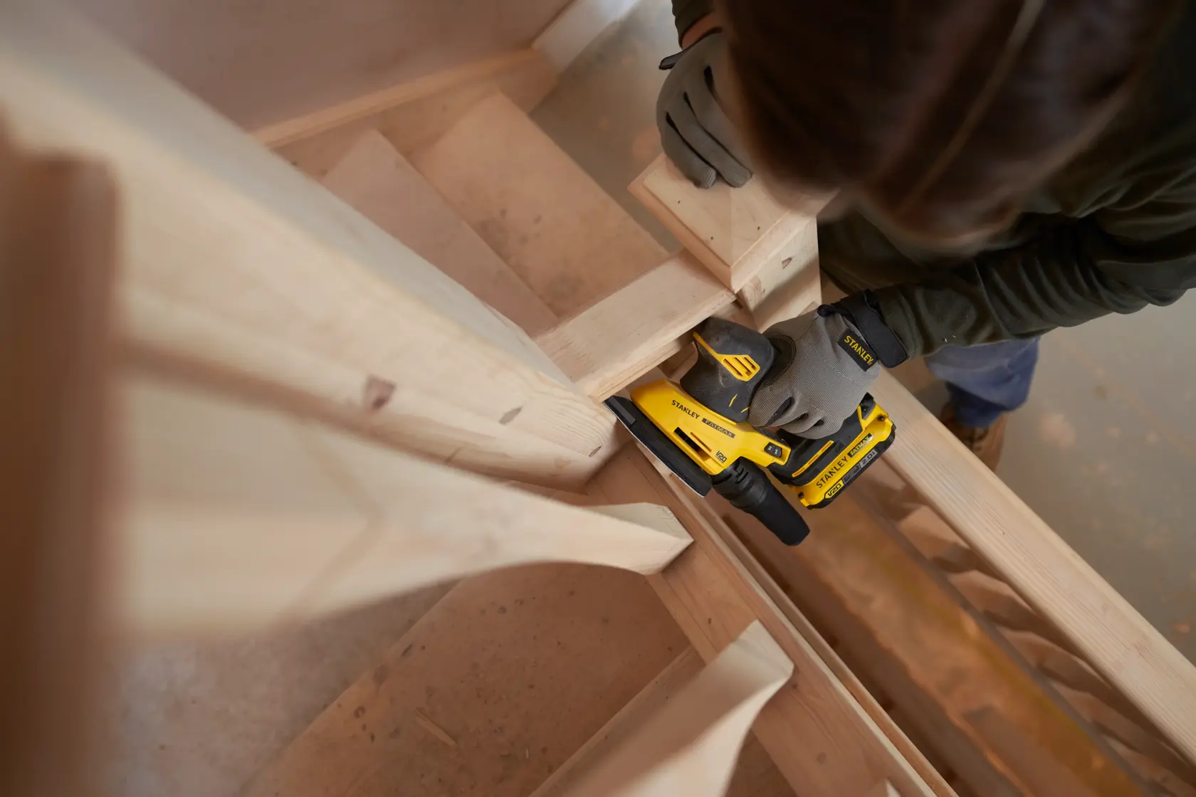 Photo of worker using STANLEY SFMCW221B sander on wooden staircase, wearing gloves, indoor woodworking setting.