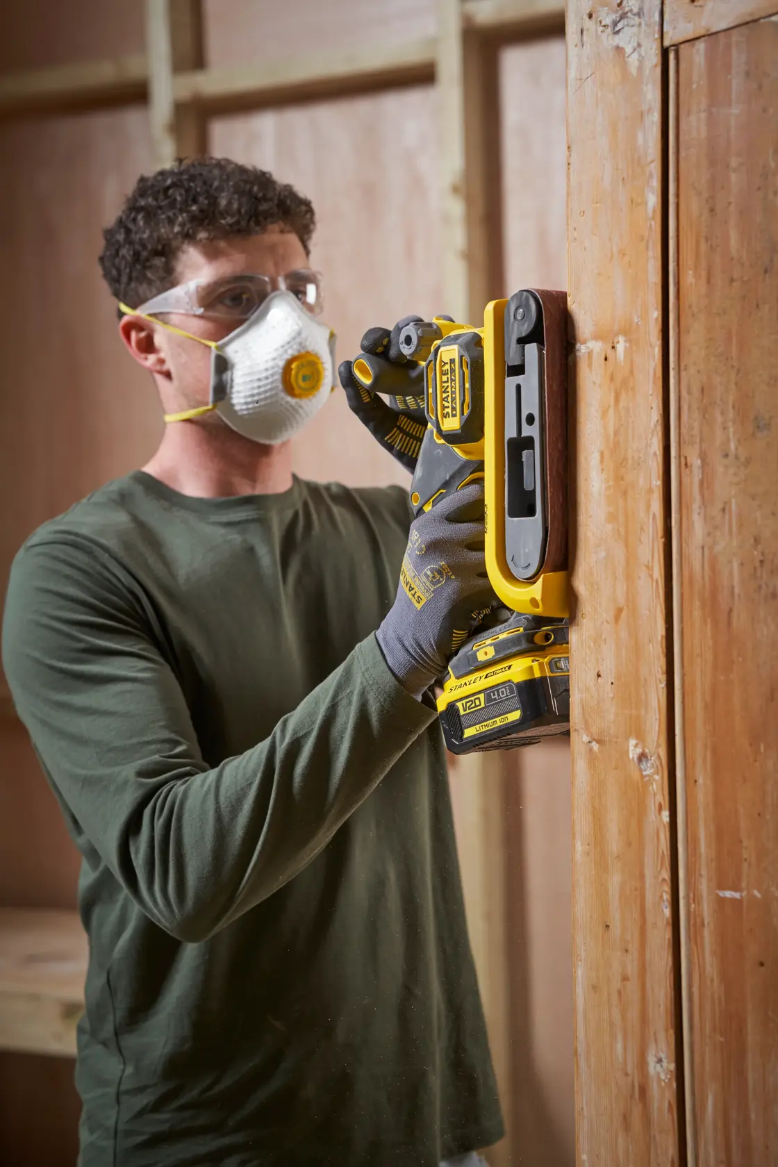 STANLEY SFMCW223B-XJ belt sander in use sanding wooden door, yellow and black tool, workshop setting.