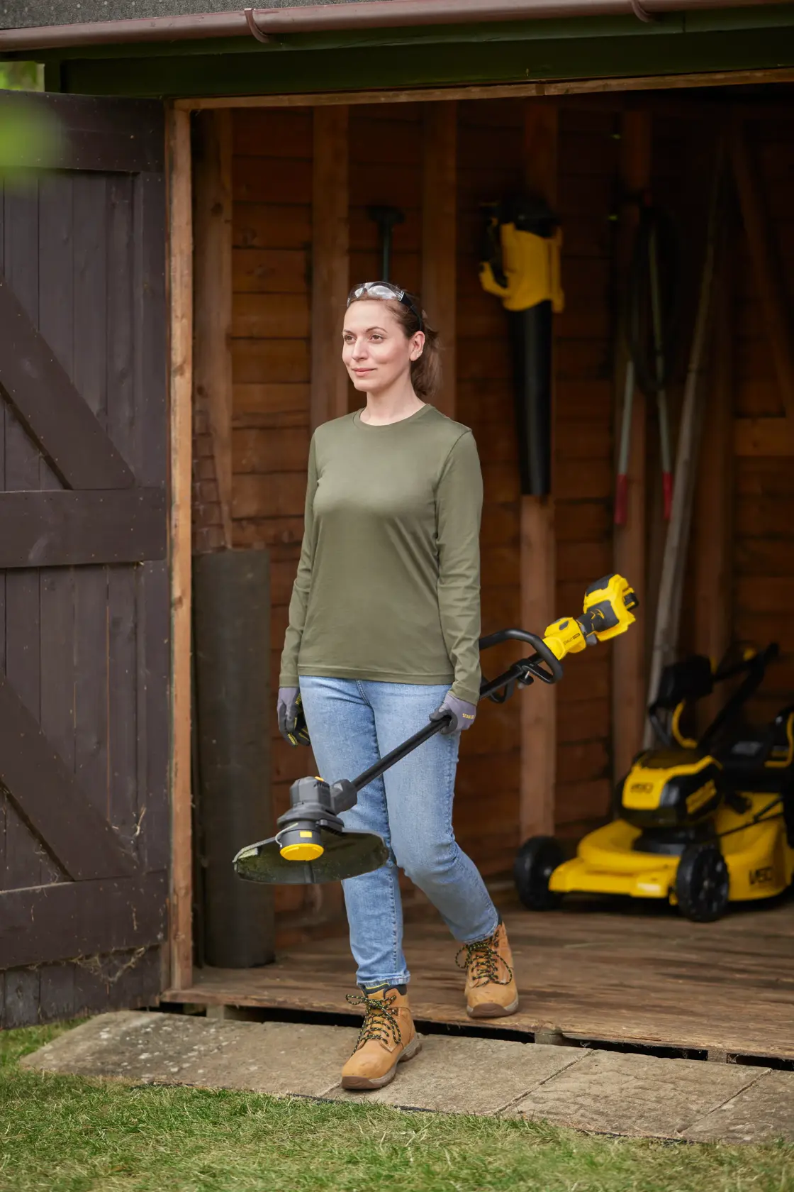 STANLEY SFMSTB930M grass trimmer held by a person in front of a shed, lawnmowers visible inside.