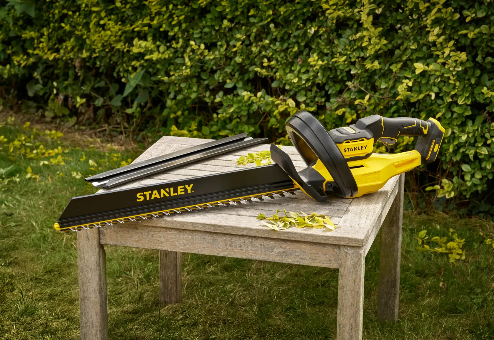 STANLEY STCHT8255B yellow and black hedge trimmer on wooden table outdoors, blades and leaves visible.
