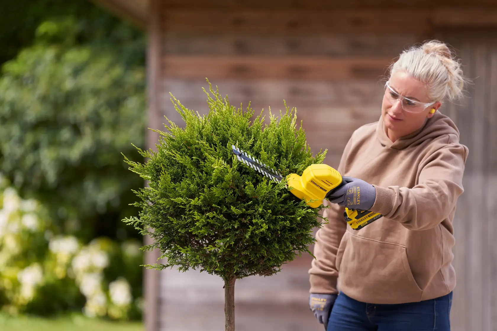 STANLEY STCSS20B hedge trimmer in use trimming a small green bush in a garden.