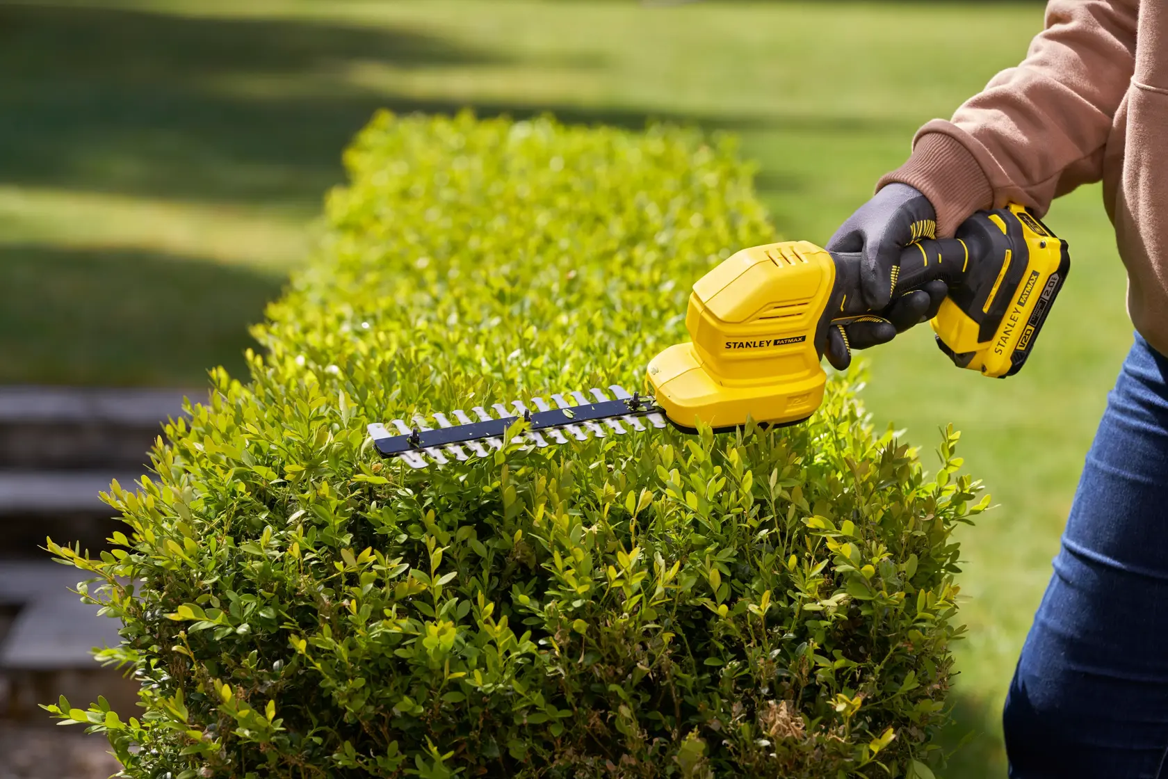 STANLEY STCSS20B cordless hedge trimmer in use trimming green bushes outdoors.