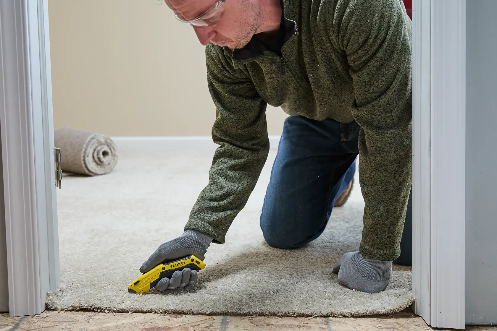 A person wearing work gloves is kneeling on a carpet and using a yellow STANLEY utility knife to cut the edge of the carpet near a doorway. A roll of carpet is visible in the background.