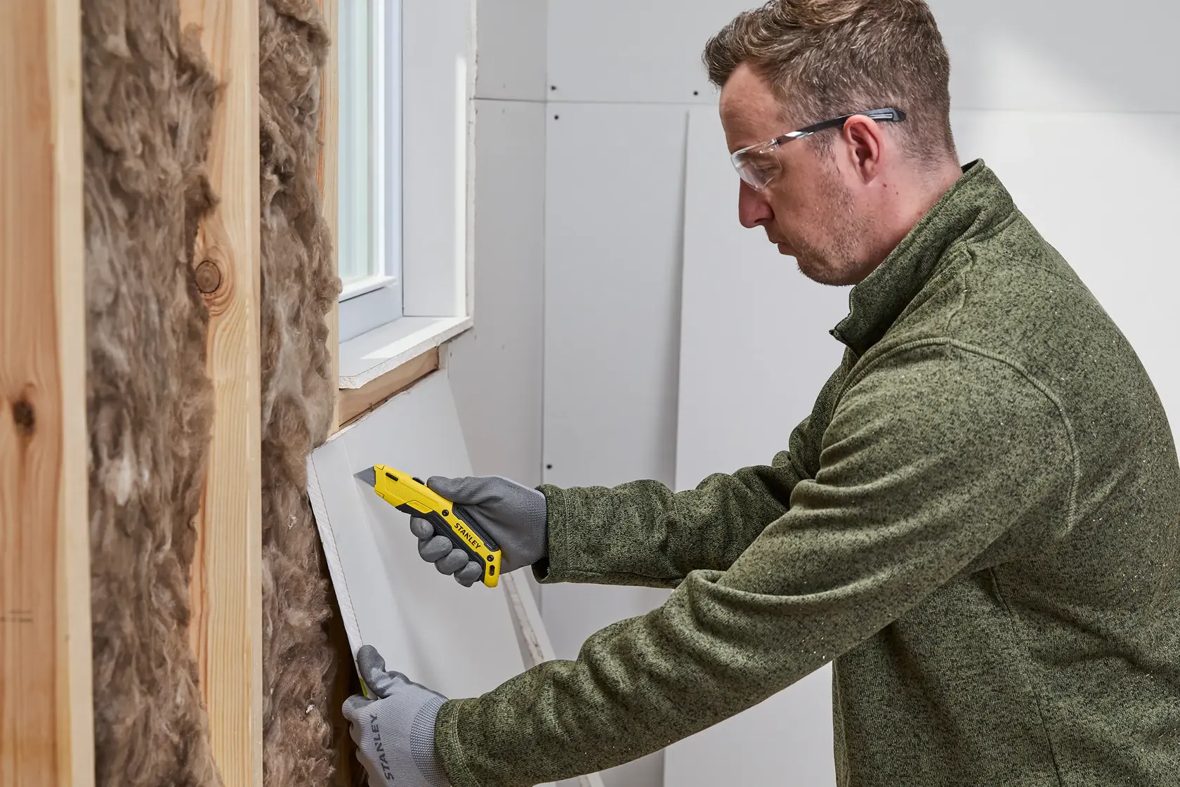 A person wearing gloves uses a yellow STANLEY utility knife to cut a piece of drywall near a window opening inside a building under construction.