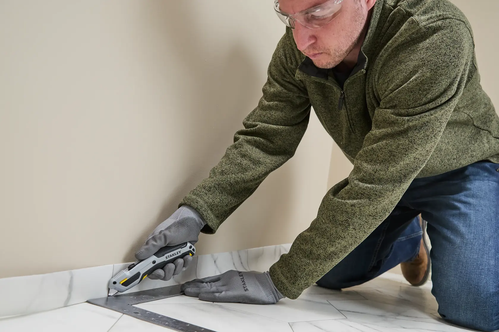 A person wearing gloves and a green jacket is using a STANLEY utility knife (model STHT10721-0) to cut along the edge of a floor near a wall, with a metal square ruler for guidance.