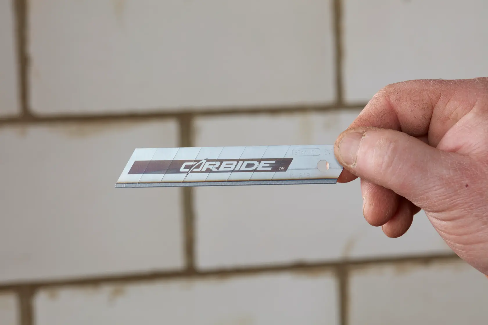 STANLEY STHT2-11818 utility knife blade, silver with carbide label, held in hand against tiled wall.