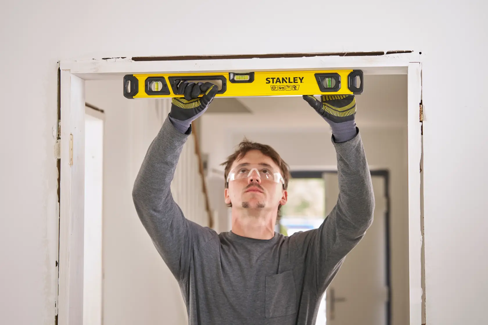 A person wearing gloves is holding a yellow Stanley spirit level against the top of a door frame in a room, checking alignment.