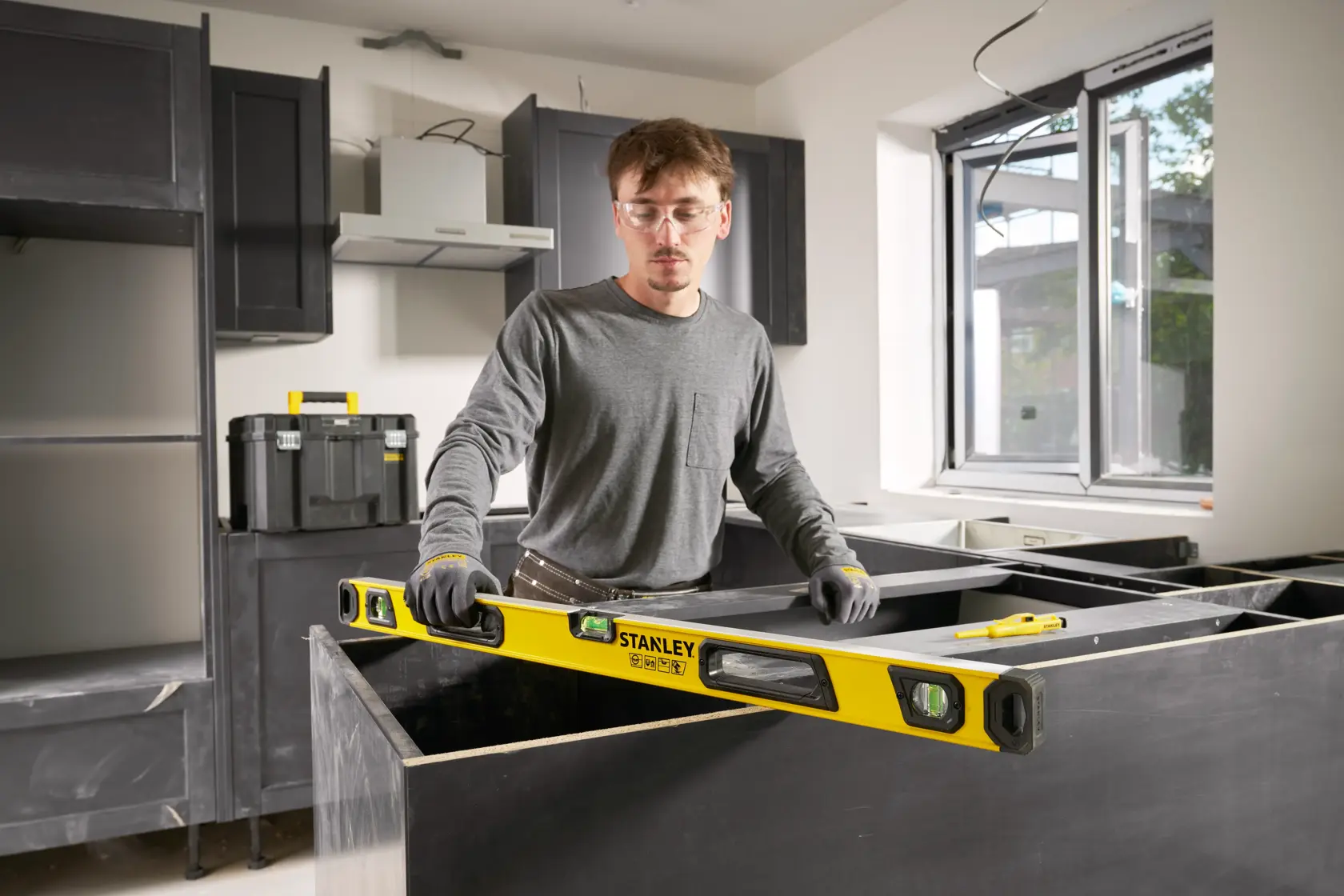 A person in work gloves is using a yellow STANLEY spirit level in a modern kitchen under construction. Cabinets, a toolbox, and a window can be seen in the background.