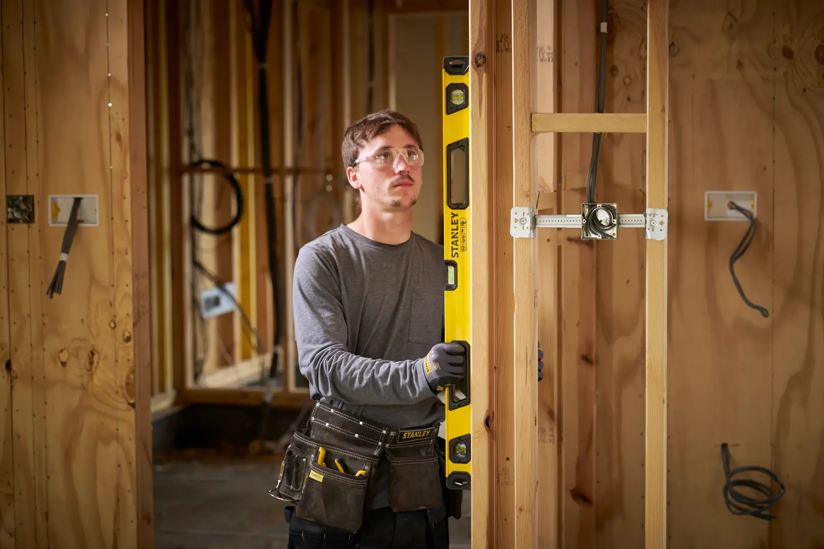 A person with a blurred face wearing a tool belt and gloves is using a yellow STANLEY spirit level to check vertical alignment on wooden framing inside a partially constructed building.