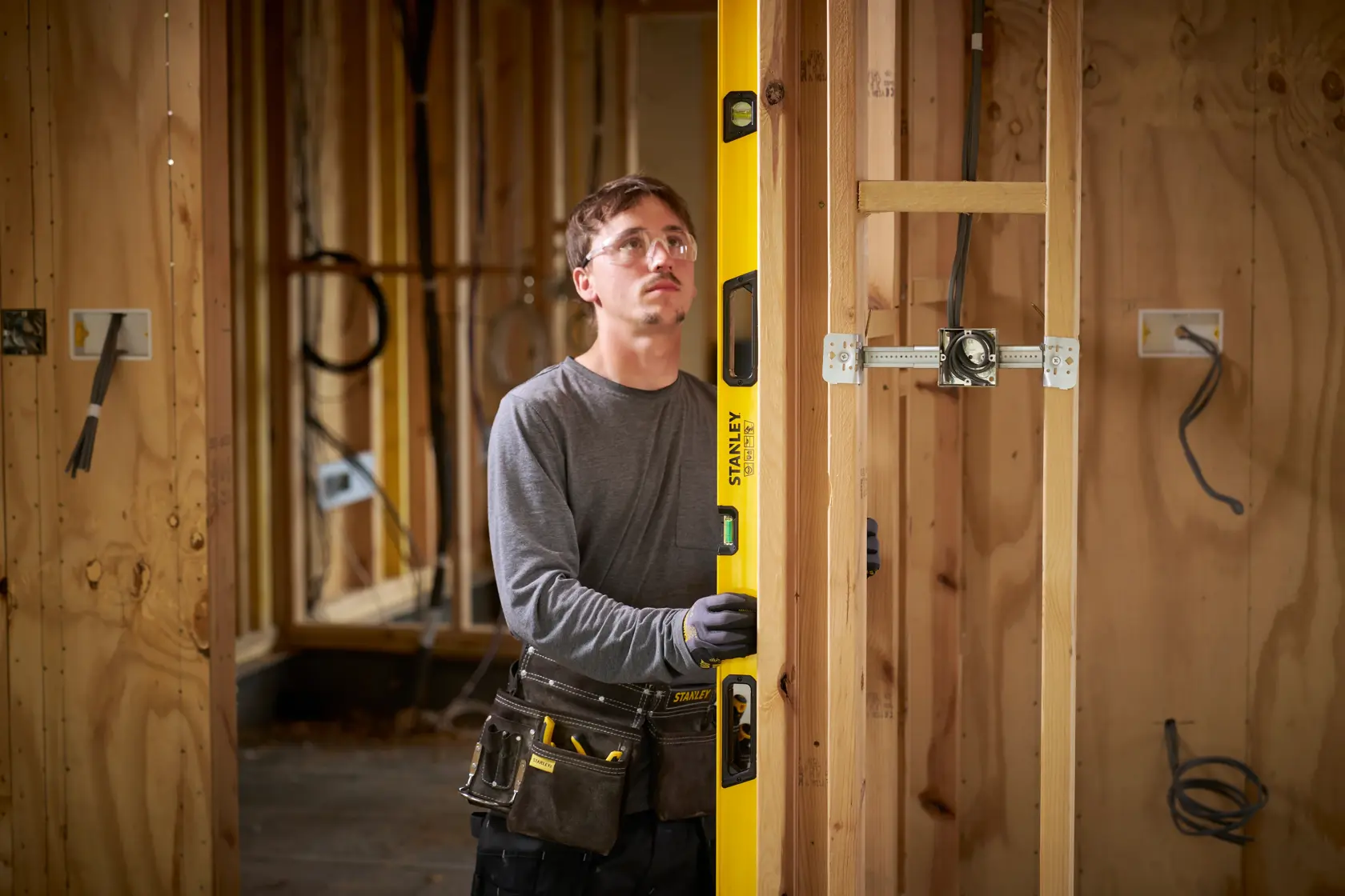 A person wearing work gloves and a tool belt is using a yellow STANLEY spirit level to check the alignment of a wooden frame at a construction site. The construction area is unfinished, with visible wooden studs and electrical wiring.