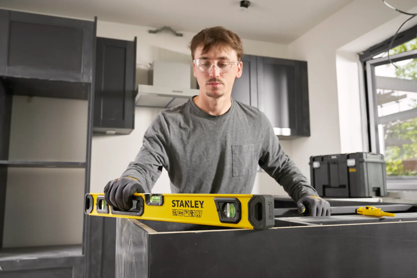 A person wearing gloves is using a STANLEY spirit level (model STHT41724-0) to check the level of a surface in a kitchen under construction. Other tools and storage boxes are visible in the background.
