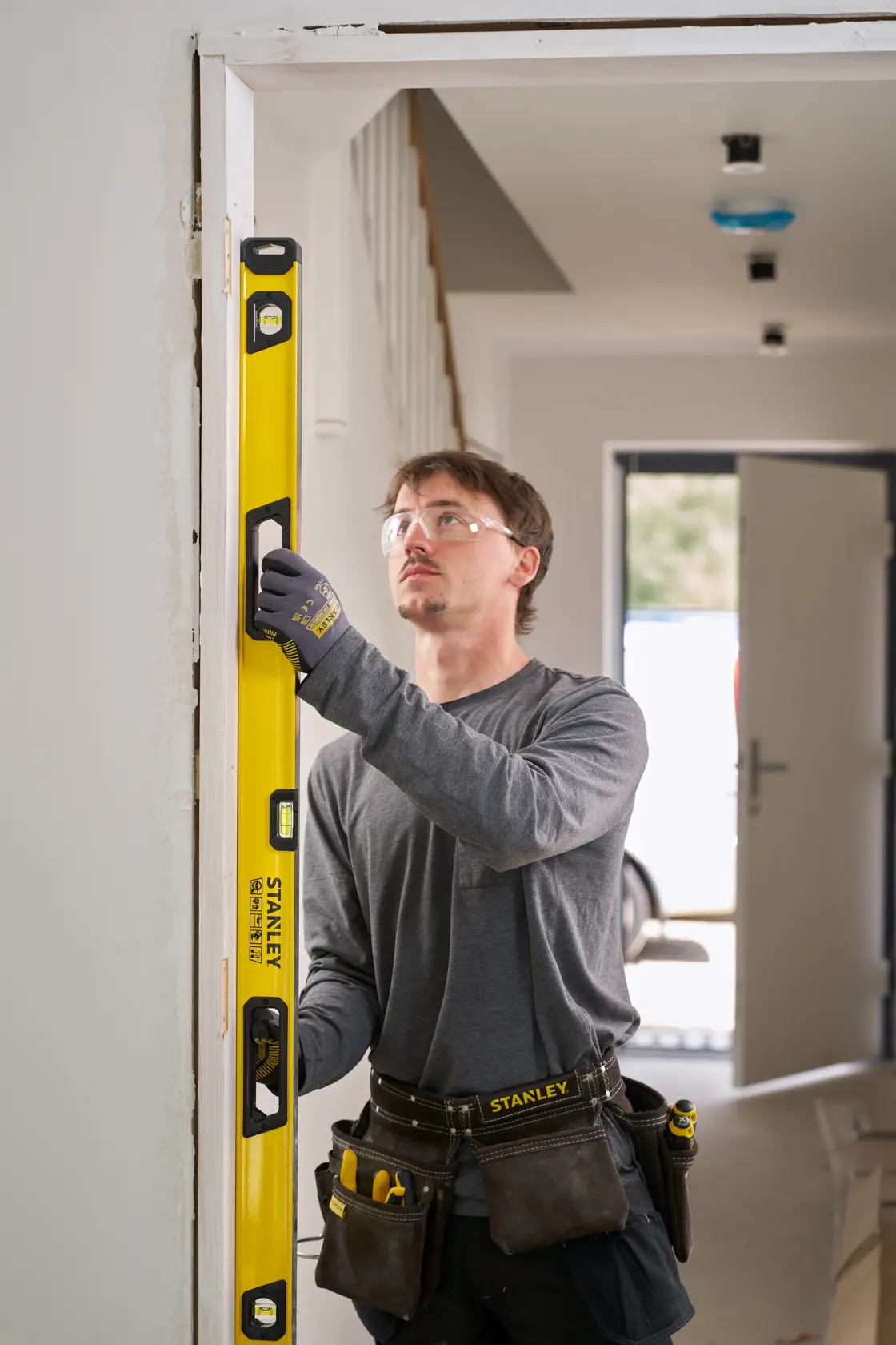 A person wearing work gloves and a tool belt is using a yellow STANLEY spirit level to check the vertical alignment of a door frame in an indoor construction setting. The person's face is blurred.