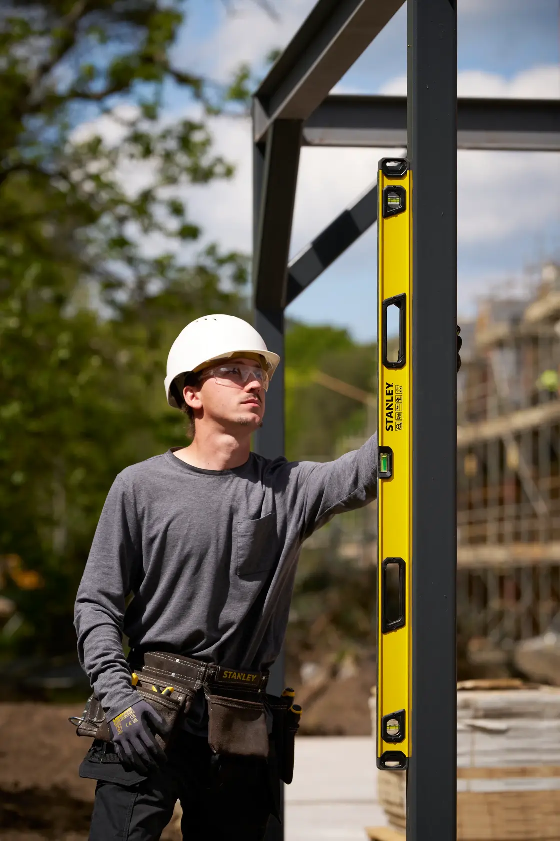 A construction worker wearing a white hard hat and a tool belt is using a yellow STANLEY spirit level against a metal frame at an outdoor construction site.
