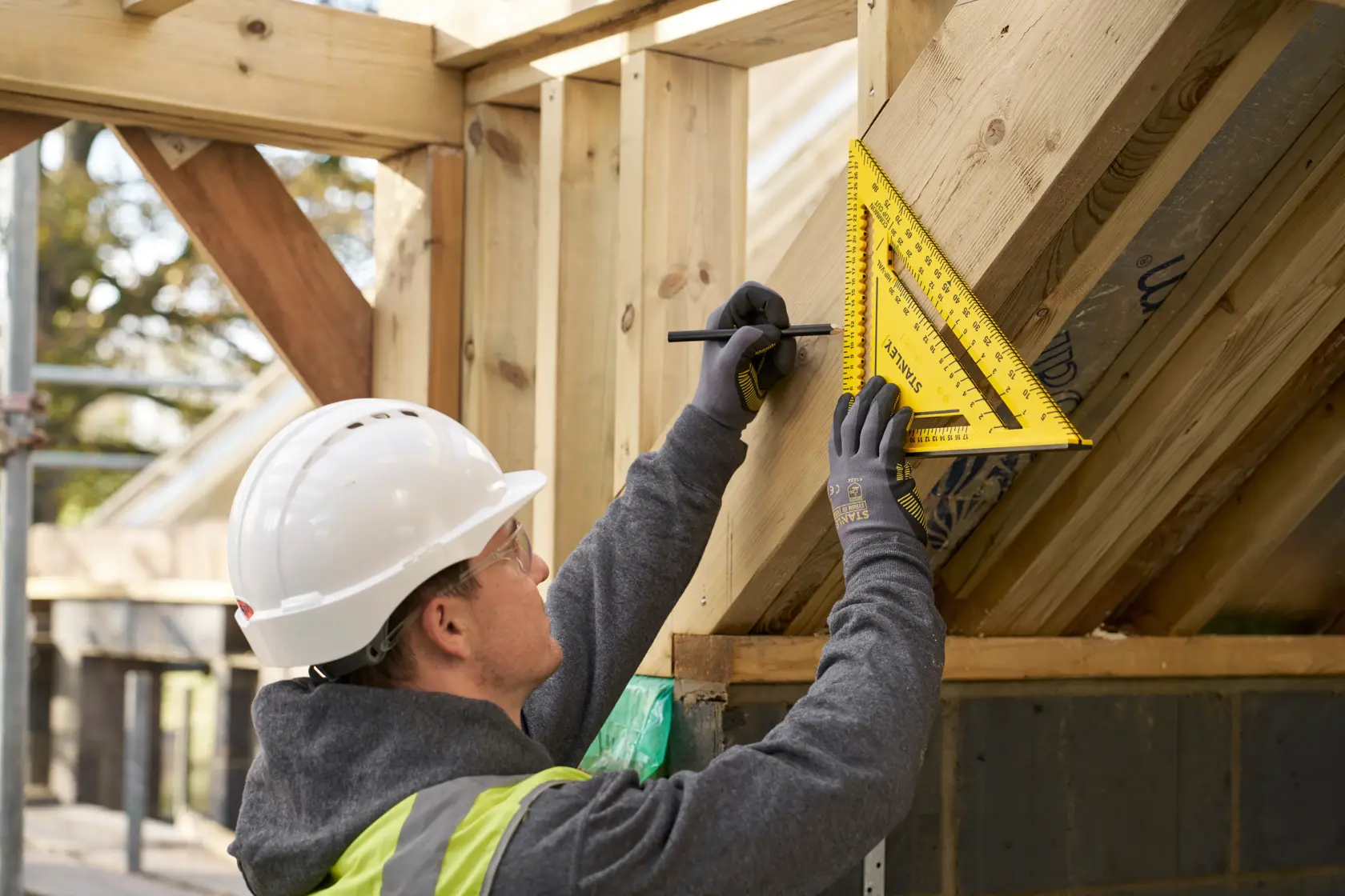 Photo of worker using STANLEY STHT46013-0 yellow rafter square on wooden roof frame, construction site background.