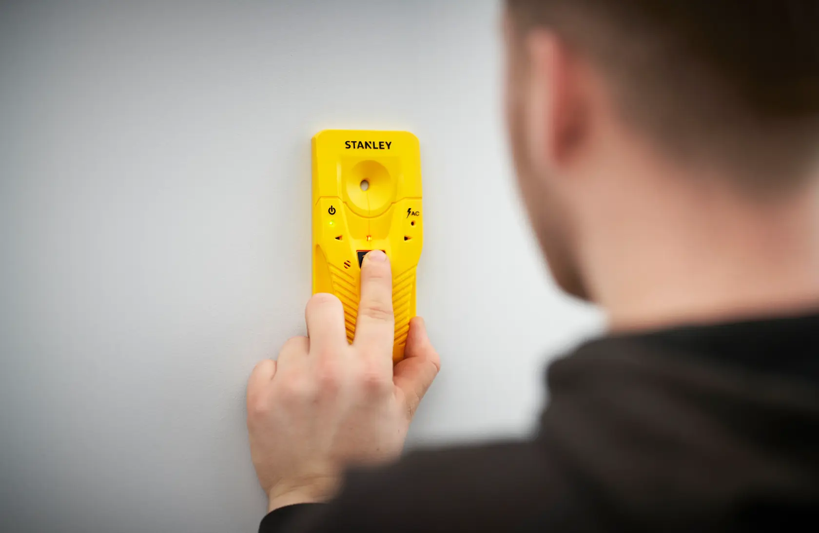 Photo of STANLEY STHT77587-0 yellow stud finder pressed against a white wall, held by a person.