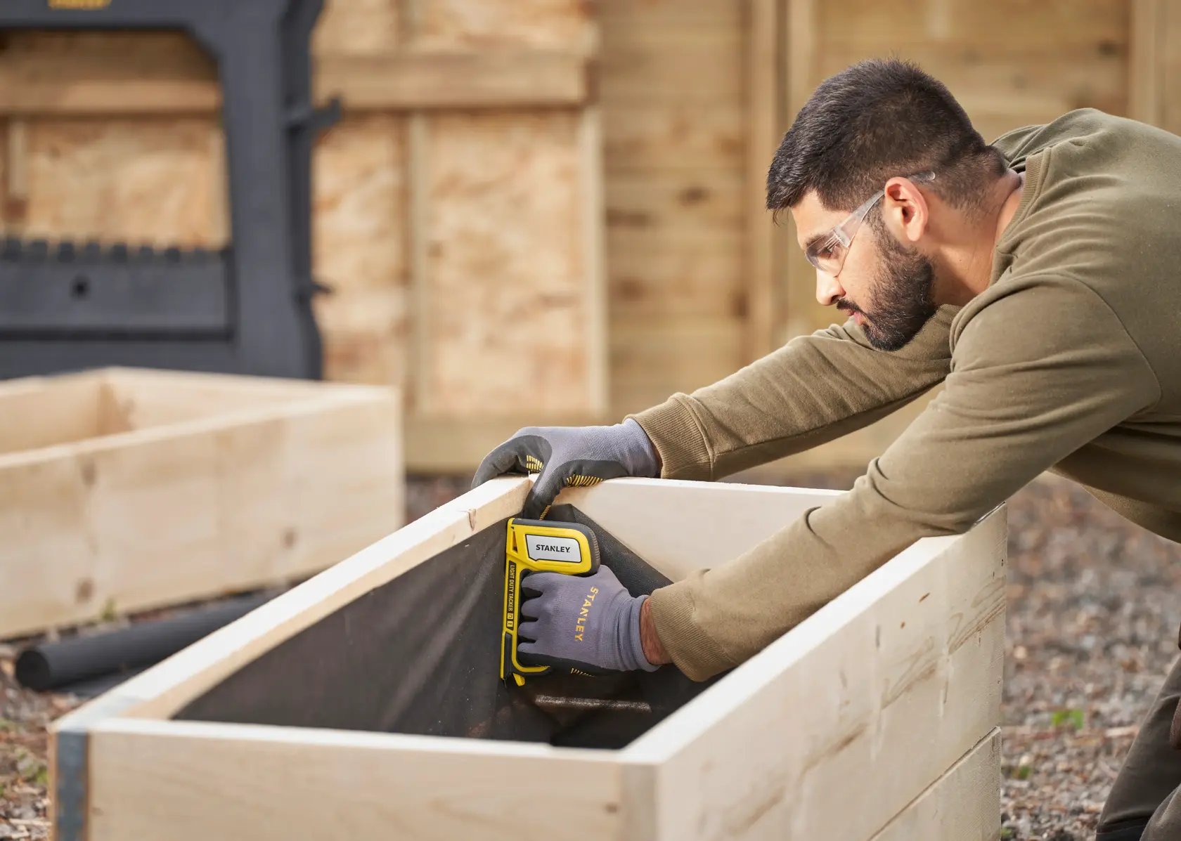STANLEY STHT70440 stapler used by a person with gloves fastening fabric inside a wooden planter box outdoors.